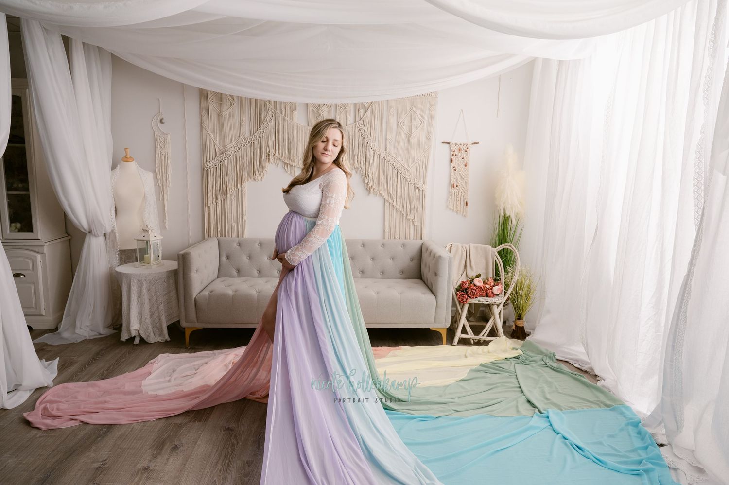 A pregnant woman stands elegantly in a flowing rainbow ombre maternity gown with soft draping fabric in a white studio setting.