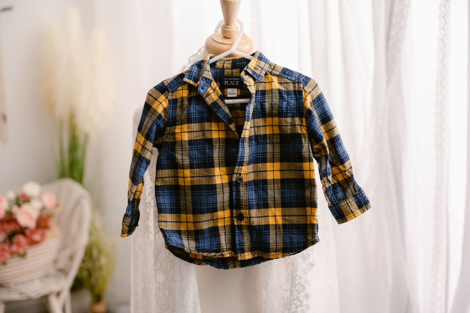 Small navy and yellow plaid flannel button-down shirt hanging from a wooden hanger against sheer white curtains.