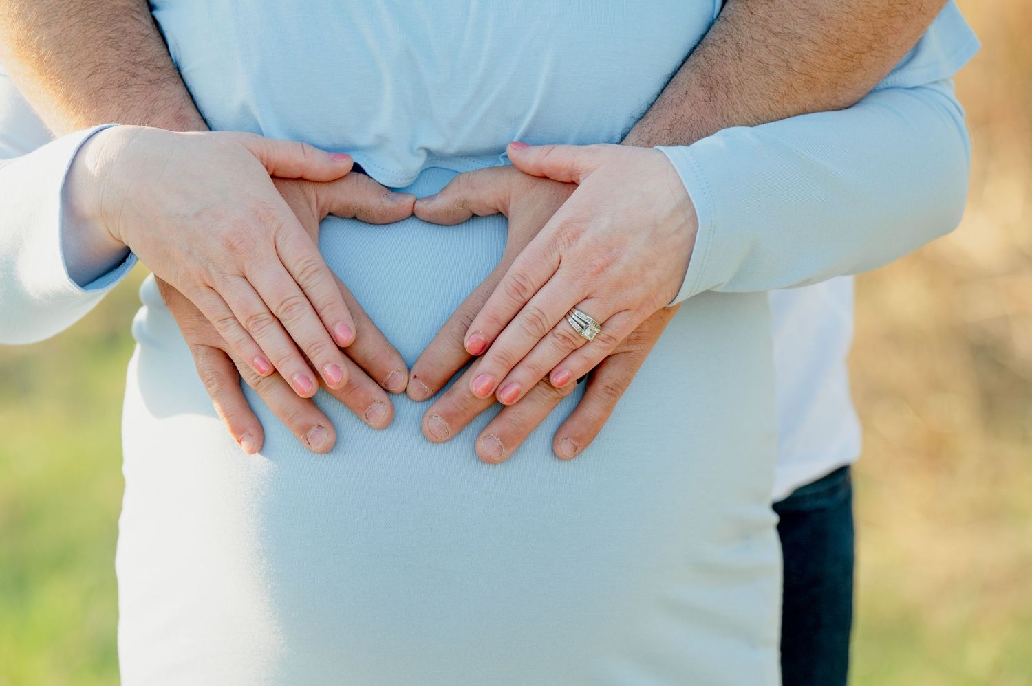 Close-up of hands forming a heart shape over a pregnant belly in light blue clothing.