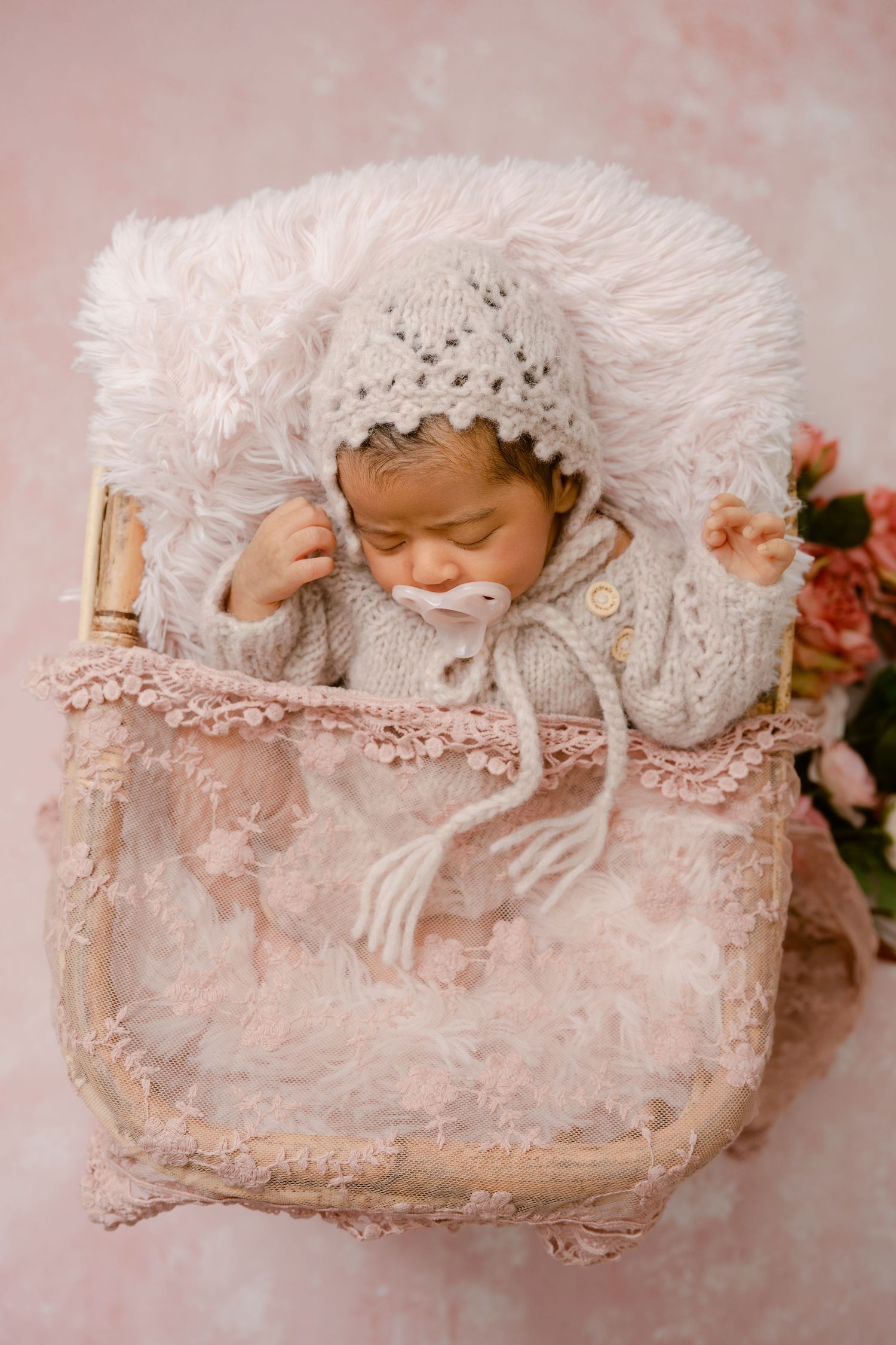 Sleeping newborn in knit bonnet nestled in pink vintage basket with lace trim.