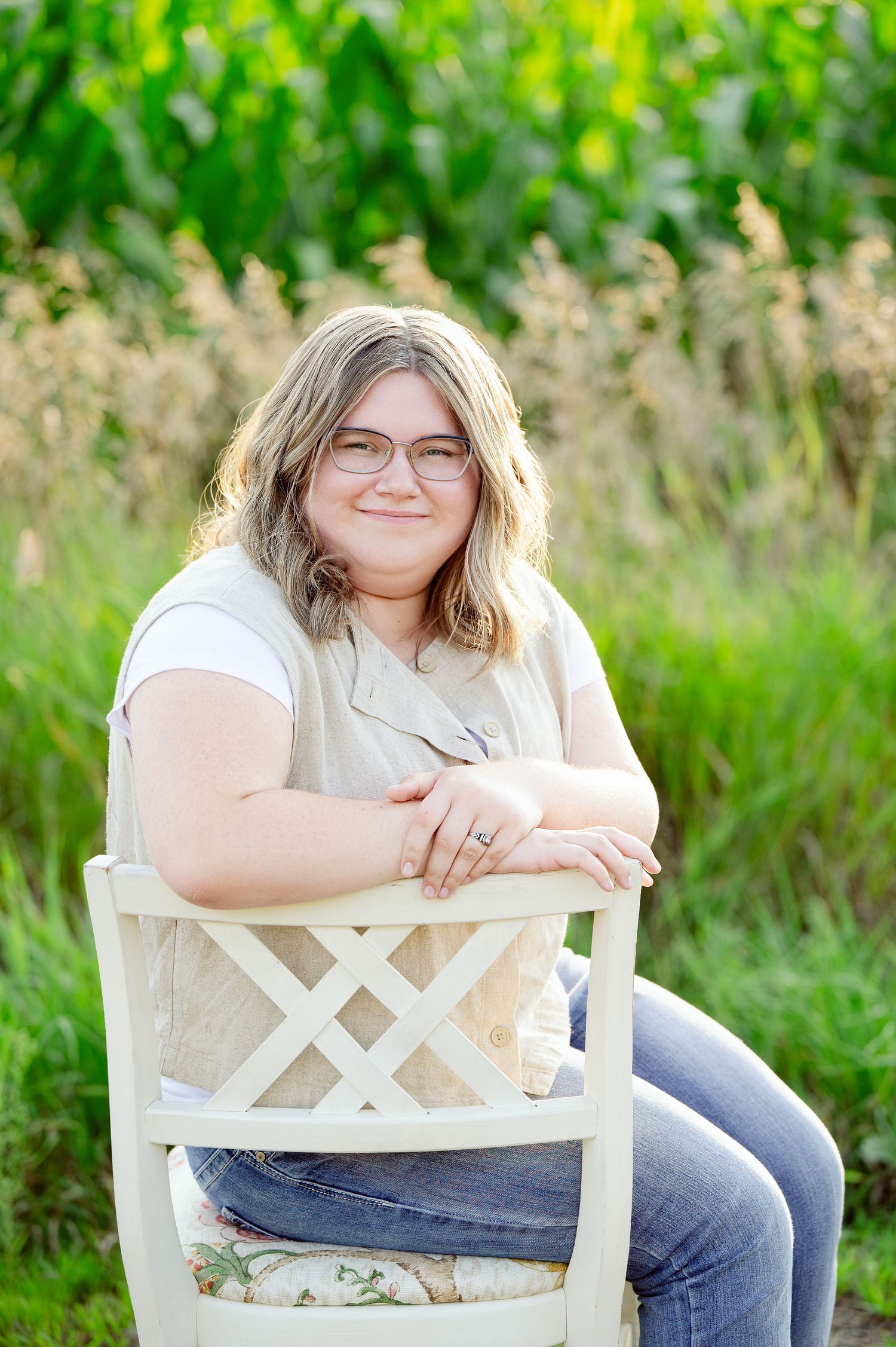 Senior Portrait in Foreston MN Person in casual attire sits on white wooden chair outdoors surrounded by greenery.