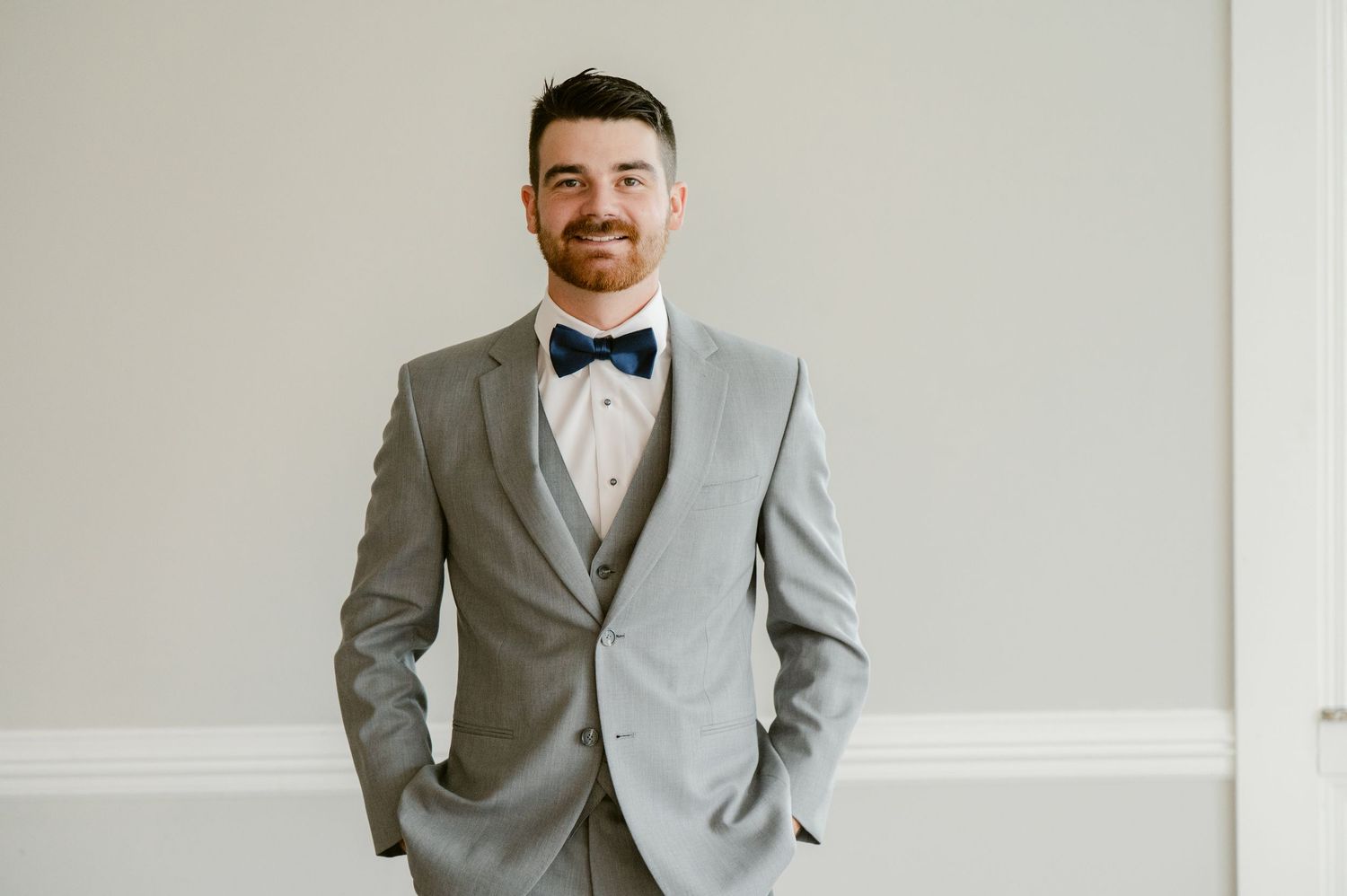 A classic portrait of someone wearing a three-piece gray suit with bowtie against a neutral wall.