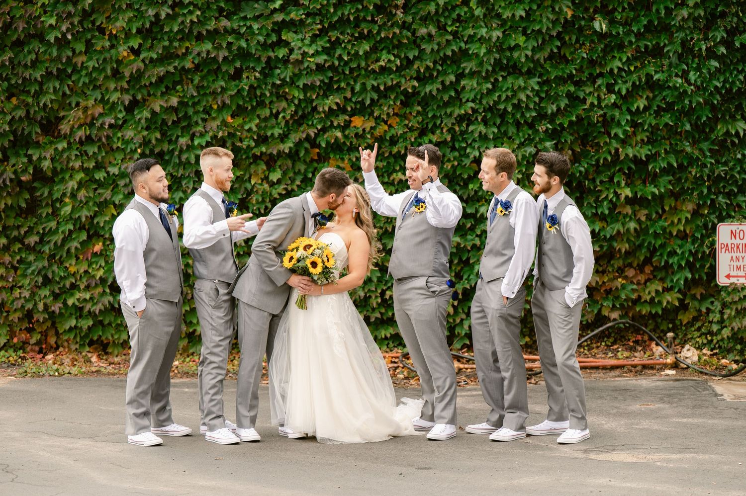 Wedding party in grey suits and sneakers celebrates with the bride against a vine-covered backdrop.