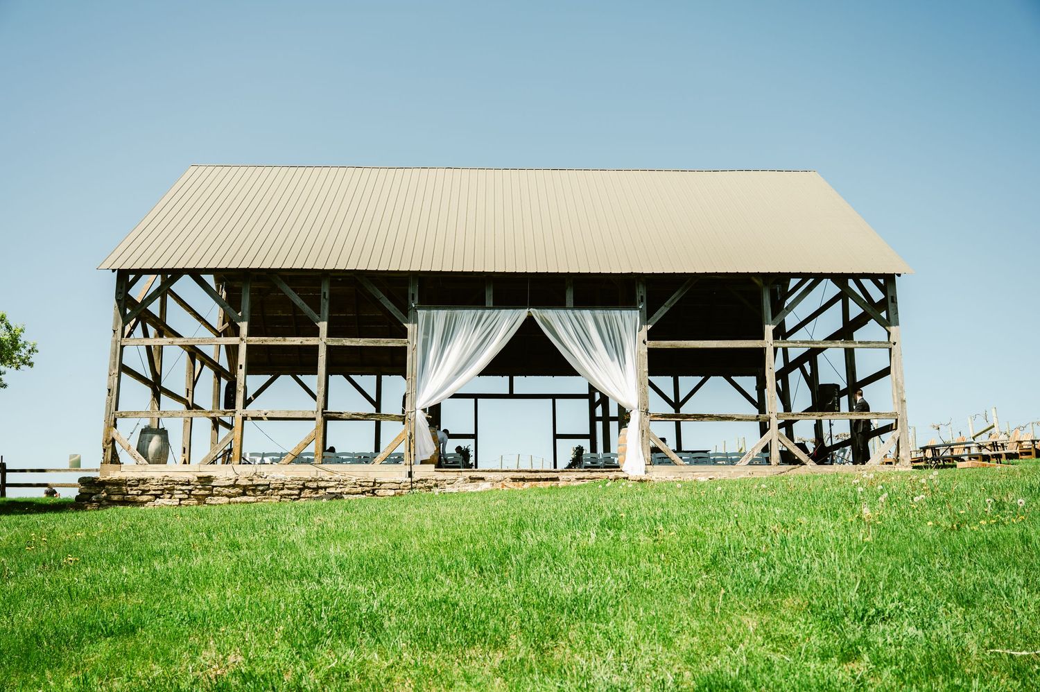 Open-air pavilion with white drapes set against blue sky on grassy hillside.