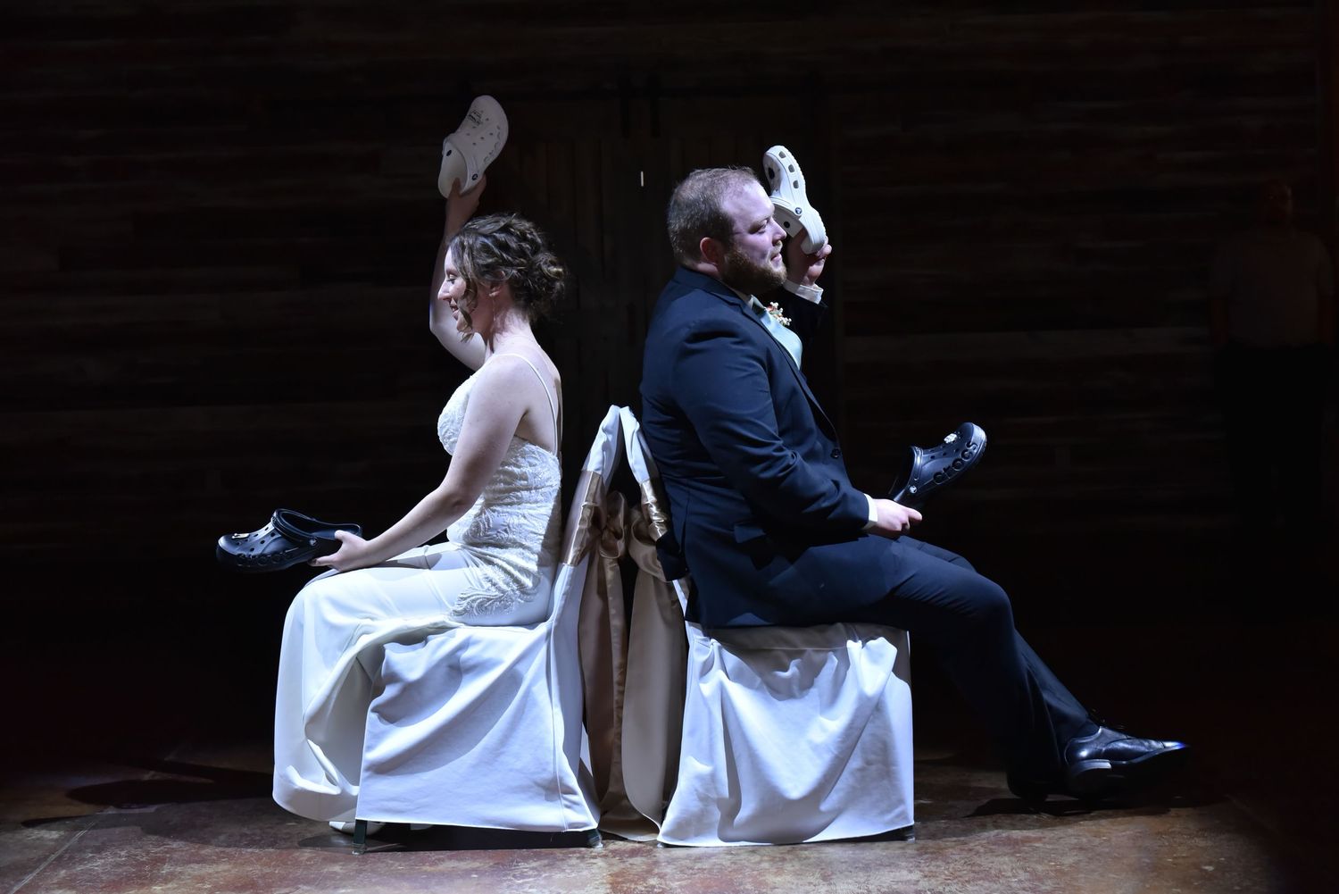 Creative wedding photo of a couple sitting back-to-back on chairs against a dark backdrop.