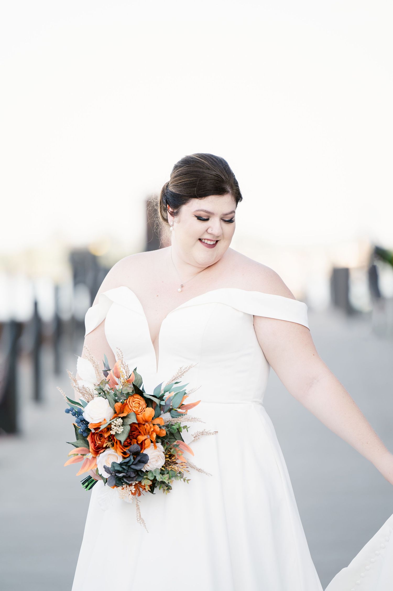 Pier B Duluth Wedding Bride in an elegant off-shoulder white wedding dress smiles while holding a vibrant orange and blue floral bouquet.