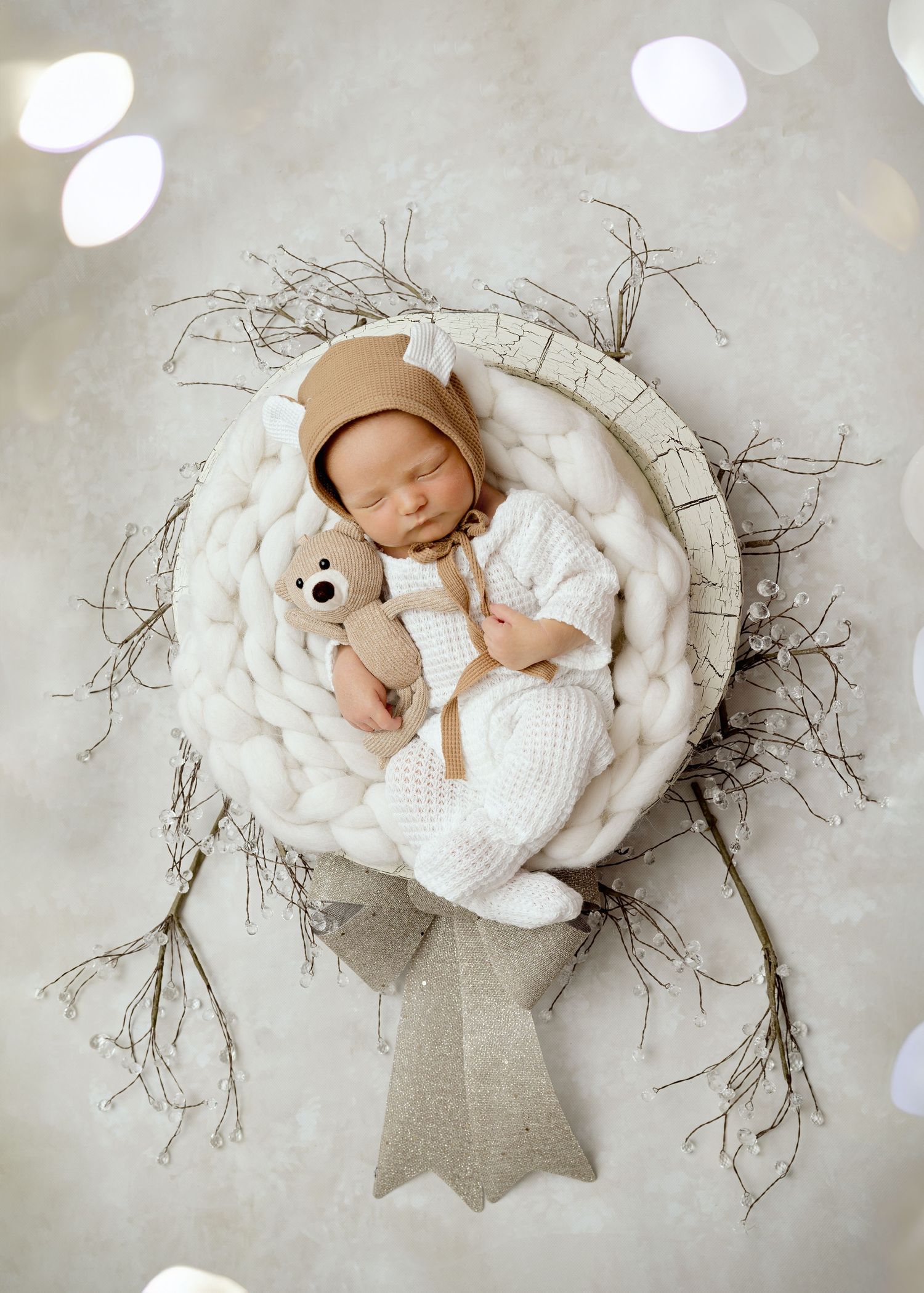 A sleeping baby wearing white clothing lies in a rustic wreath basket with a plush teddy bear on soft white fabric.