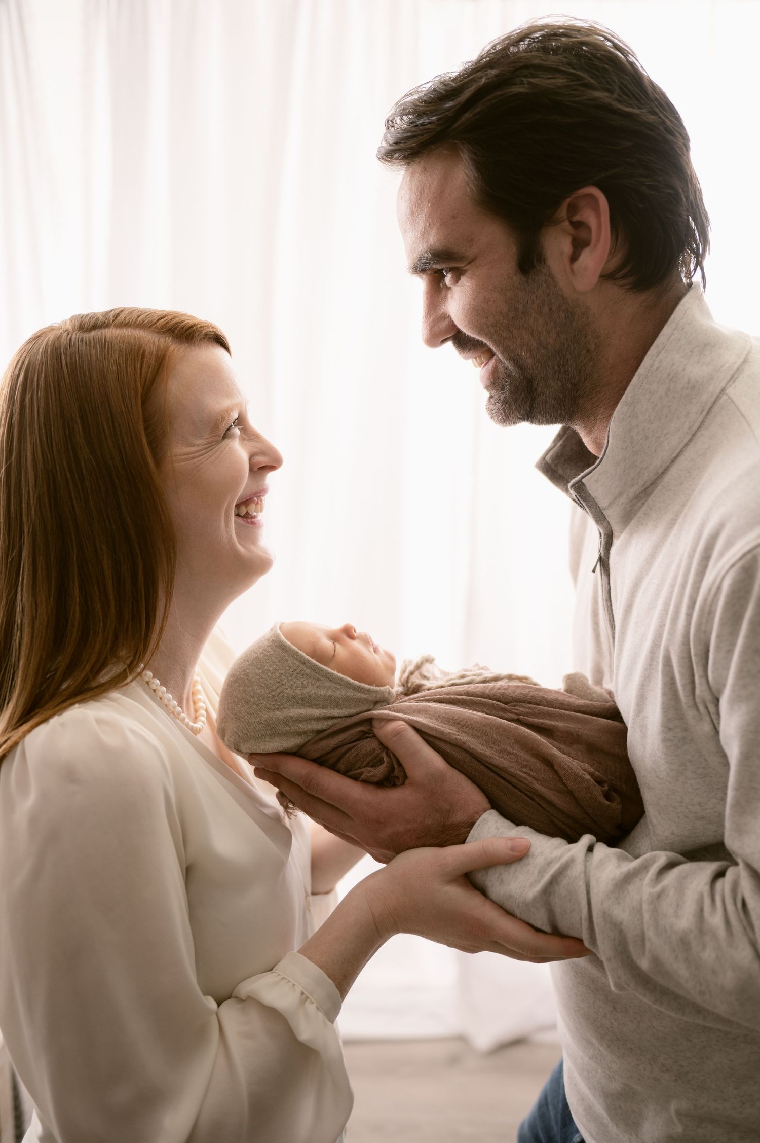 A couple shares a joyful moment while holding their swaddled newborn baby near white curtains.