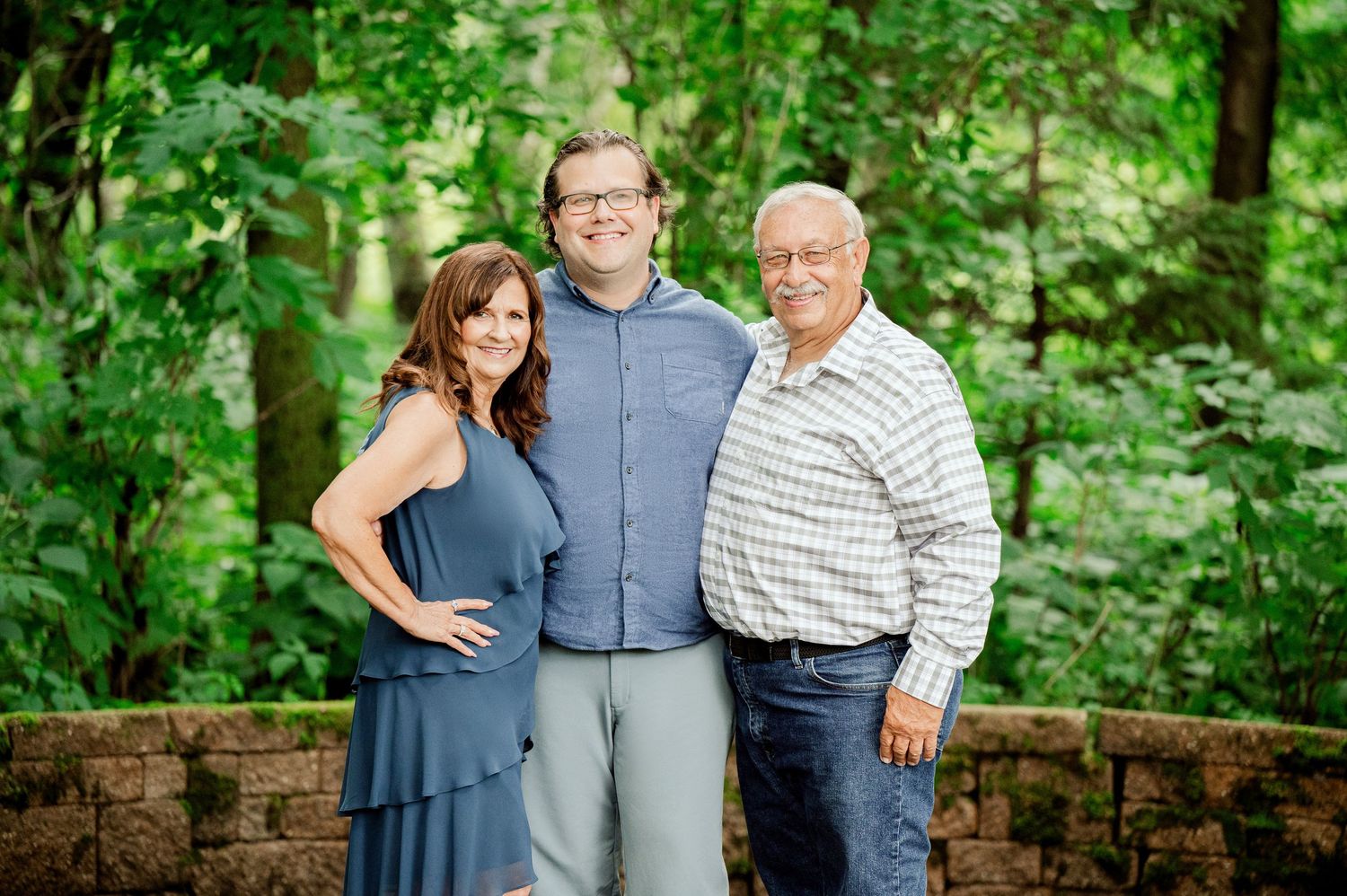 Clear Lake MN Family Photos: Three people pose together in a lush green forest setting with stone wall visible.