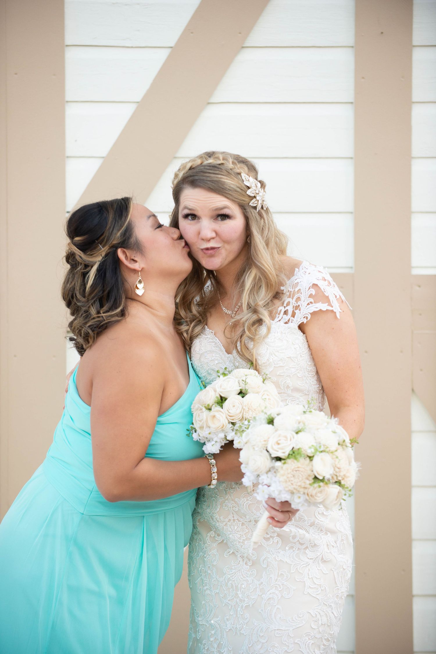 A bridesmaid in turquoise dress shares a moment with bride wearing lace gown against white wall. Milaca Minnesota Weddings