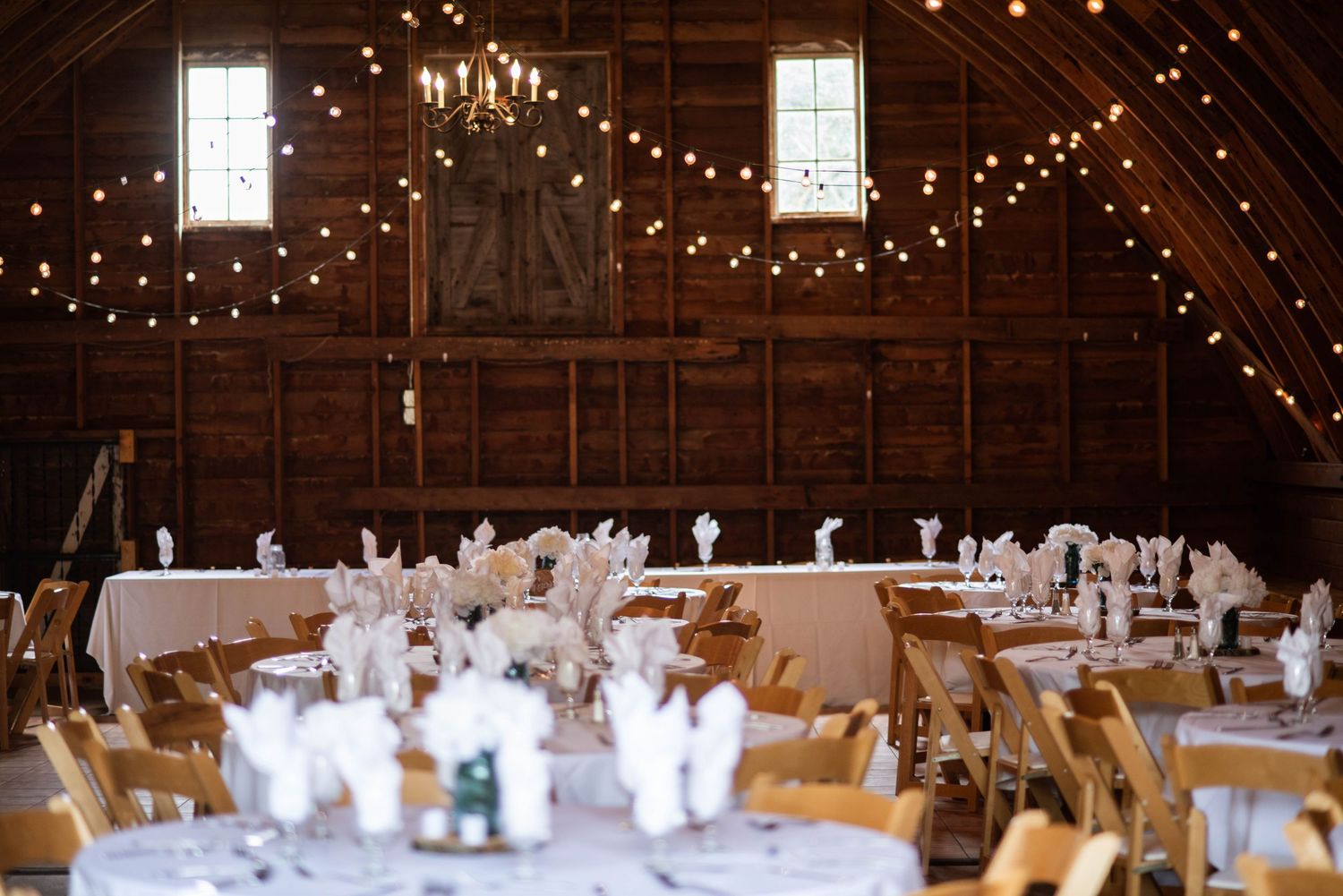 A rustic barn wedding reception setup with twinkling lights and round tables decorated in white linens.
