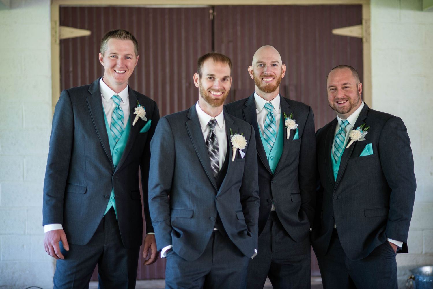 Groomsmen in black suits with turquoise accents stand against neutral wall for formal portrait.
