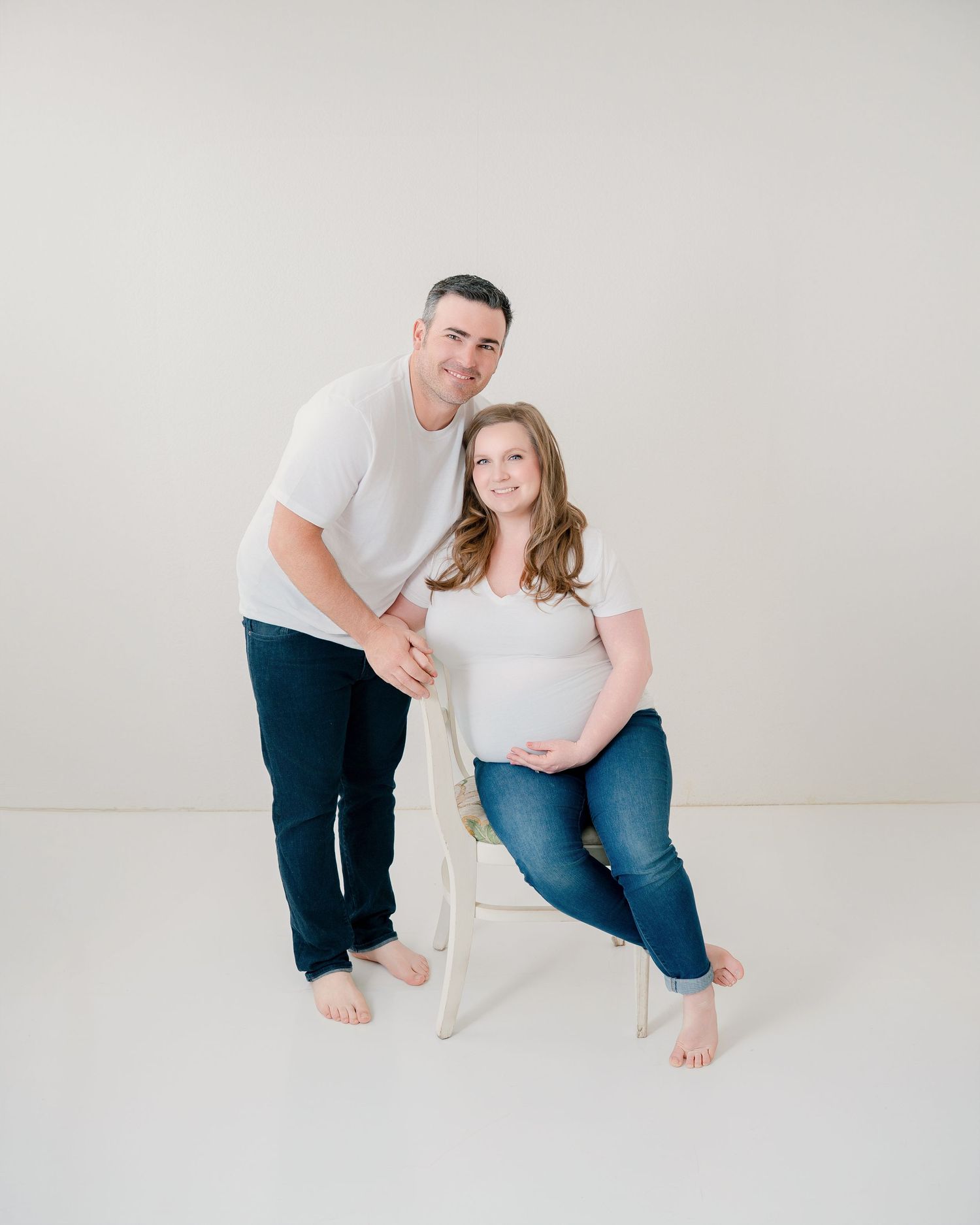 Couple in matching white shirts and dark jeans poses casually with barefoot on white backdrop.