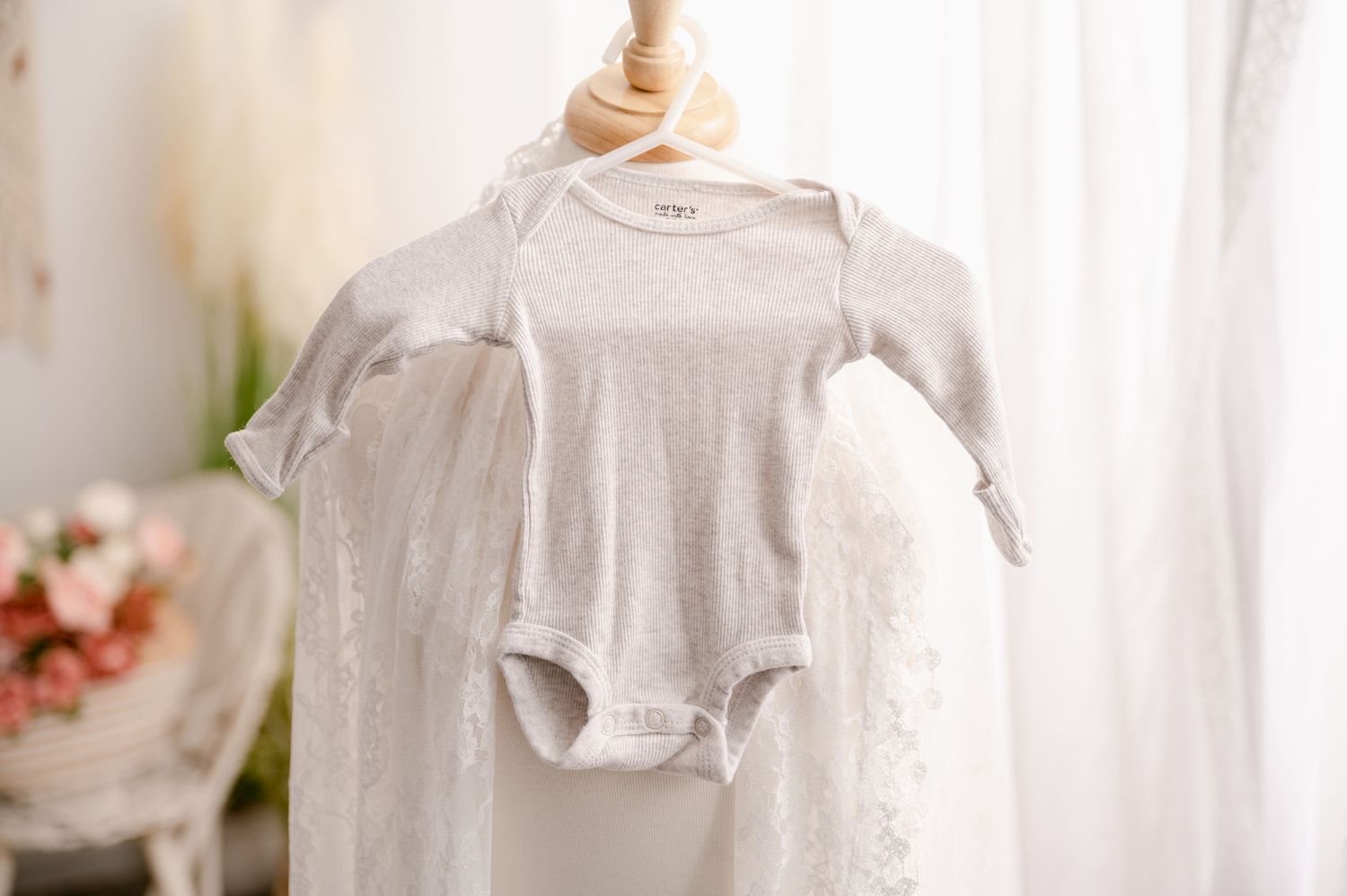 A small gray baby onesie hangs on a white wooden hanger against a light background with pink flowers visible.