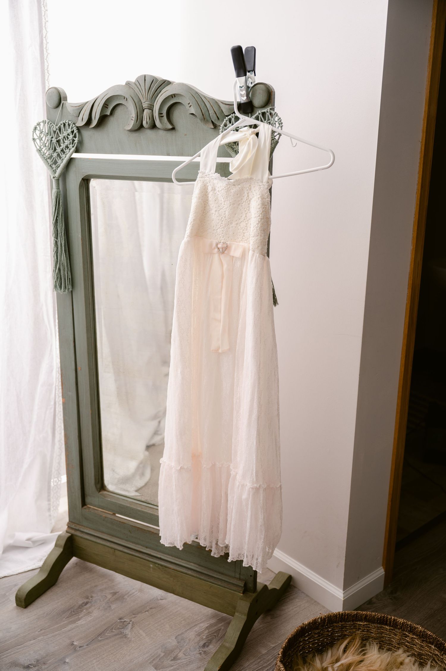 A vintage-style white lace dress hangs on a hanger beside an ornate sage green standing mirror.