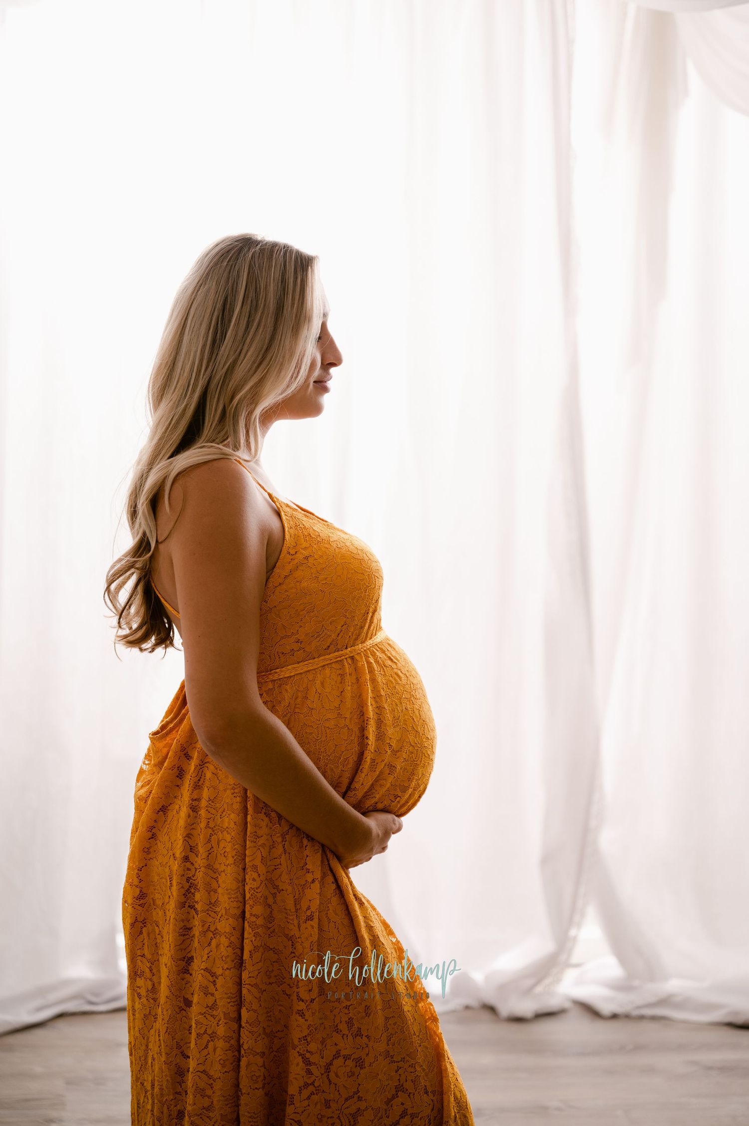 A side profile view of an expectant mother in a mustard yellow lace dress standing by sheer white curtains.