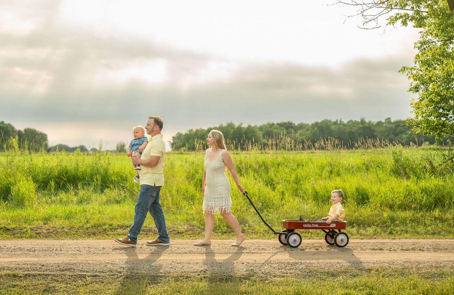 Family walking on a dirt path in summer with a red wagon through peaceful green countryside under cloudy skies.