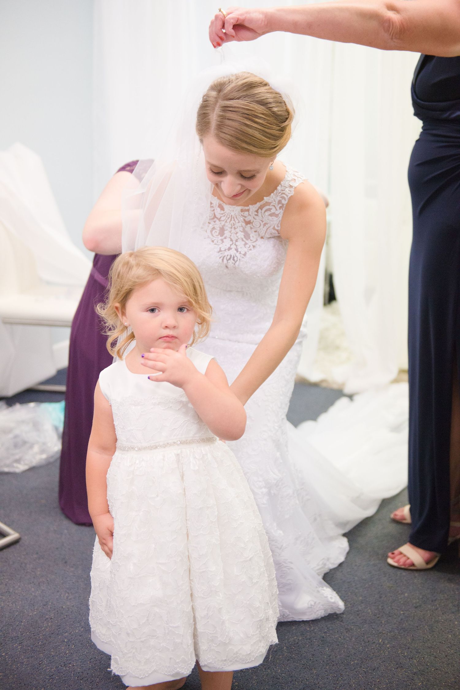A bride in a lace gown kneels beside a young flower girl in a white dress during wedding preparations. Andover wedding at Magestic Oaks Golf