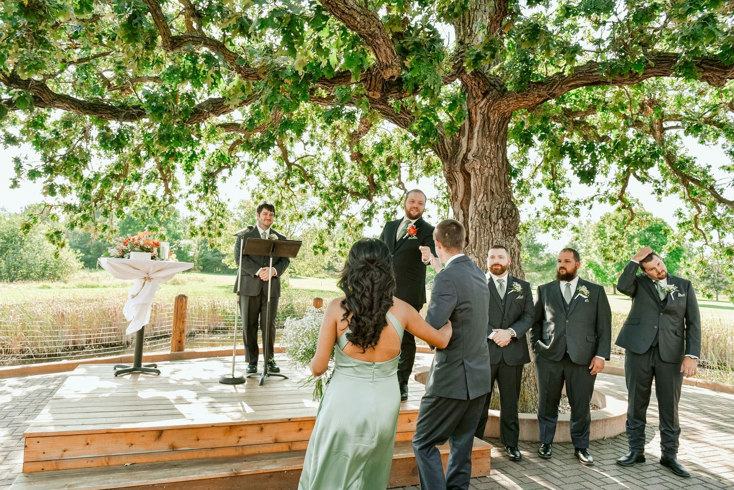 The Grands At Mulligans. A wedding party gathers on a wooden deck under the shade of a large tree during an outdoor ceremony.