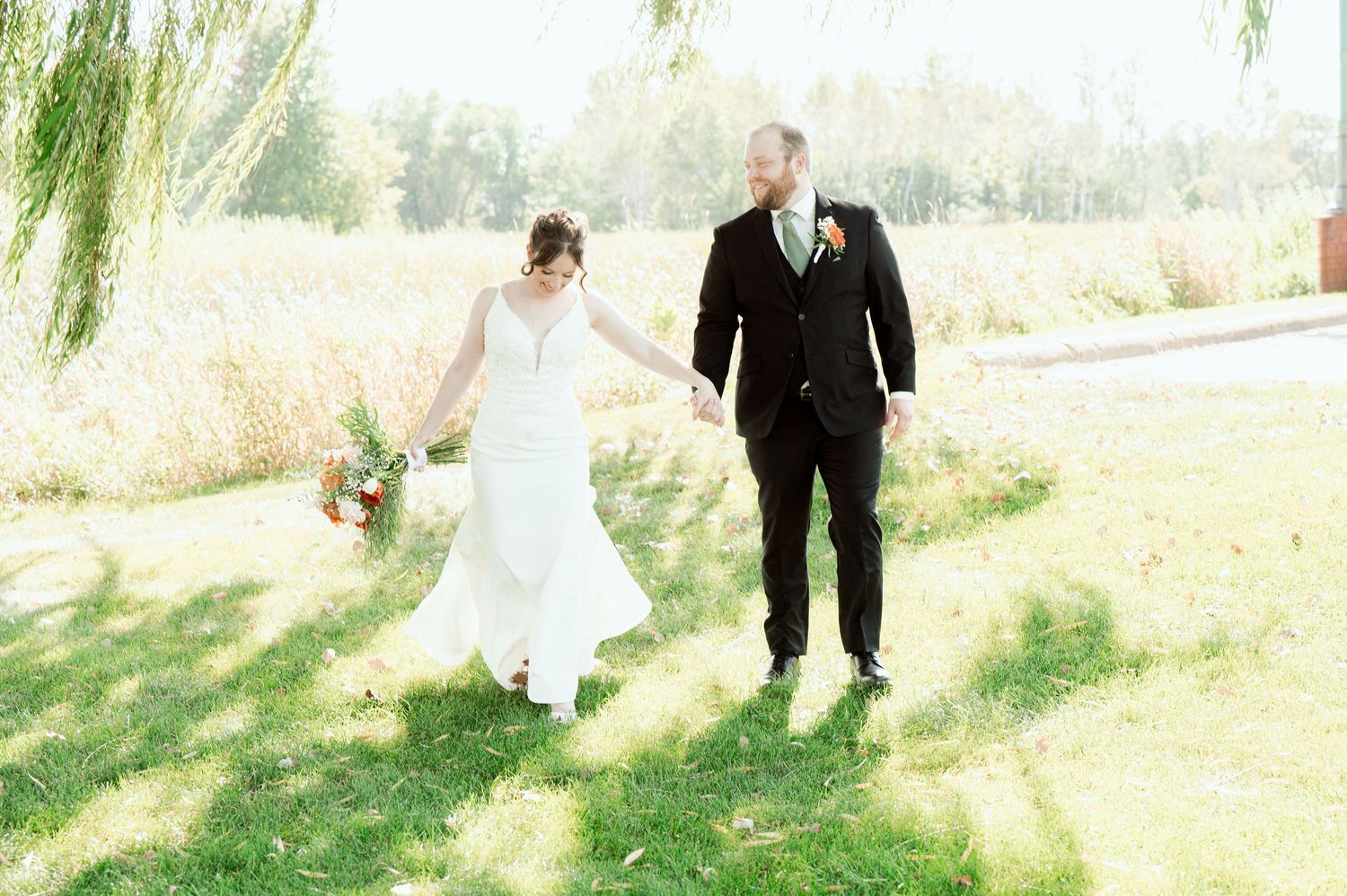 A bride and groom walk together under a willow tree during their outdoor wedding.