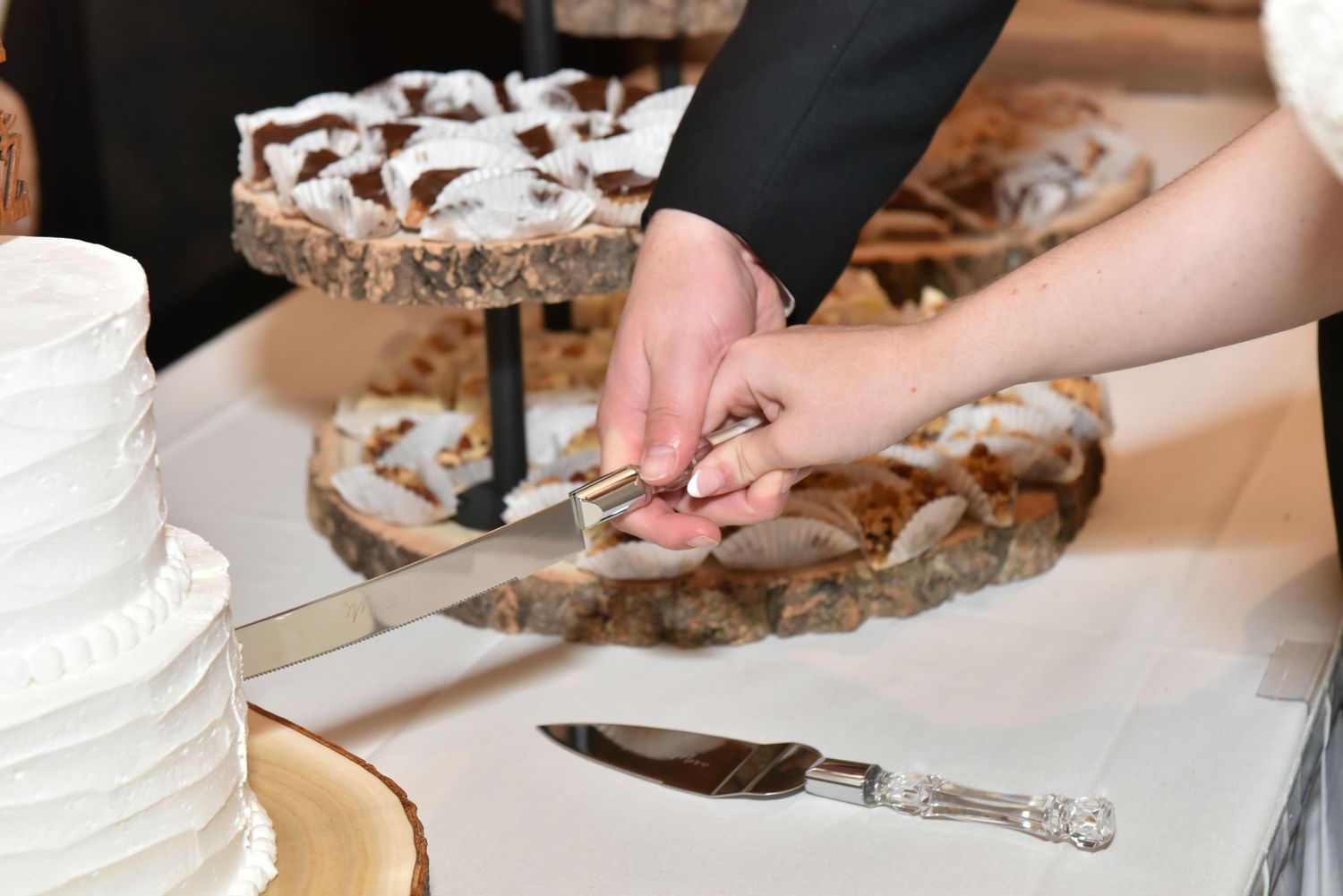 Close-up of hands cutting into a white tiered wedding cake at reception.