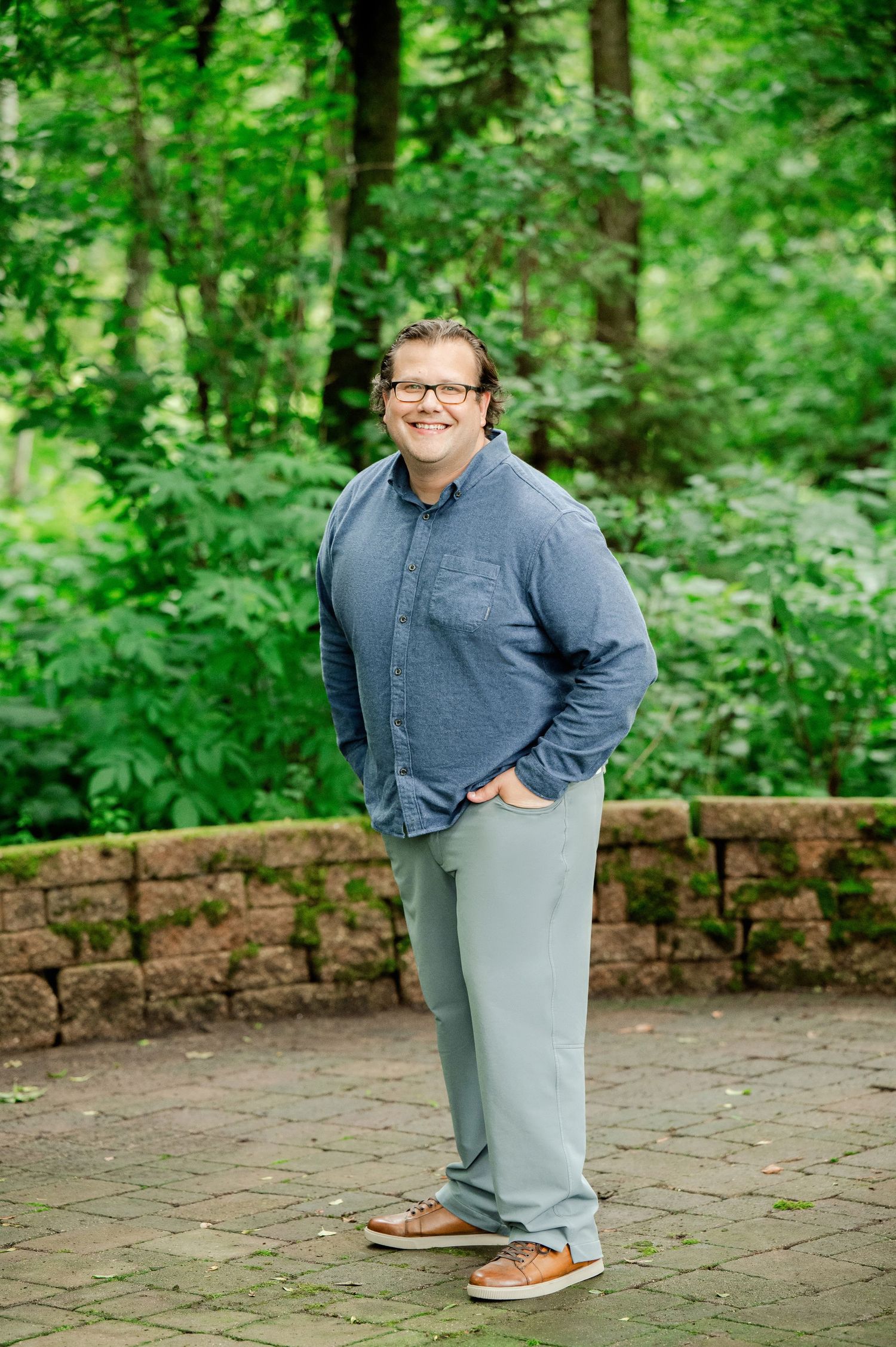 A person wearing glasses stands smiling on a brick paver patio surrounded by lush green forest scenery.