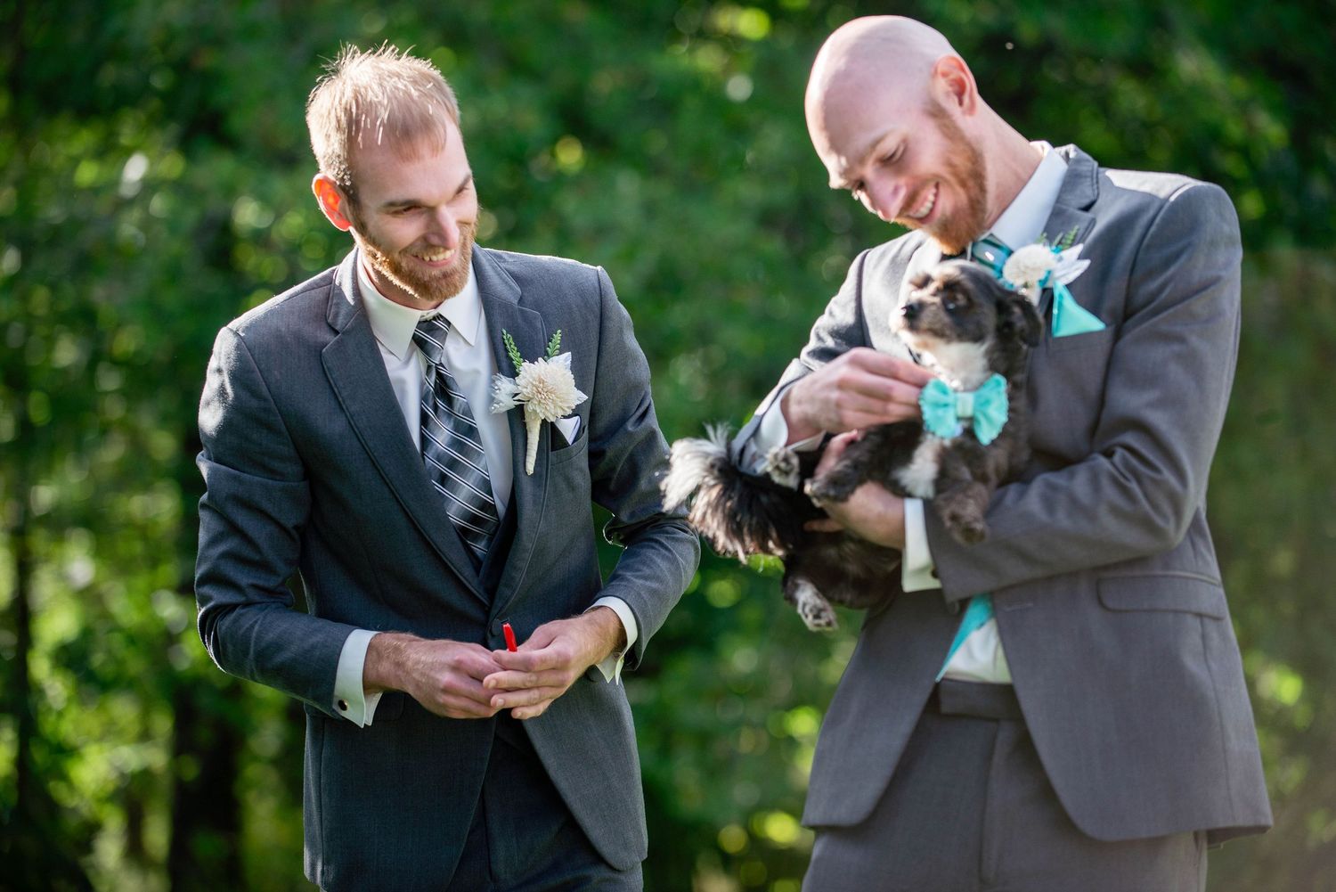 Two groomsmen in suits share a moment with a small dog wearing turquoise at an outdoor wedding. Rum River Barn Milaca Minnesota Weddings
