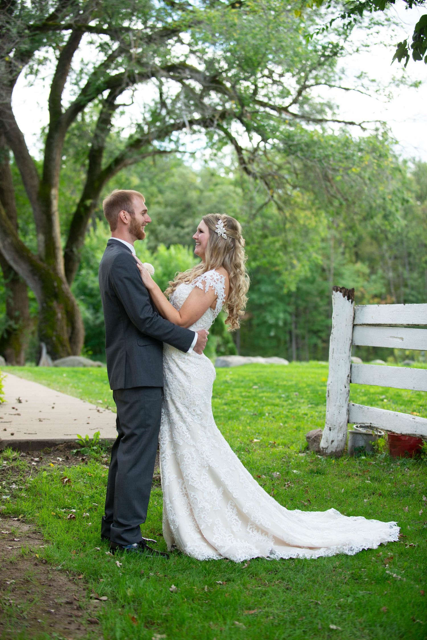 A romantic wedding portrait of newlyweds embracing on a lush green lawn with a white fence in the background. Rum River Barn Milaca Minnesot