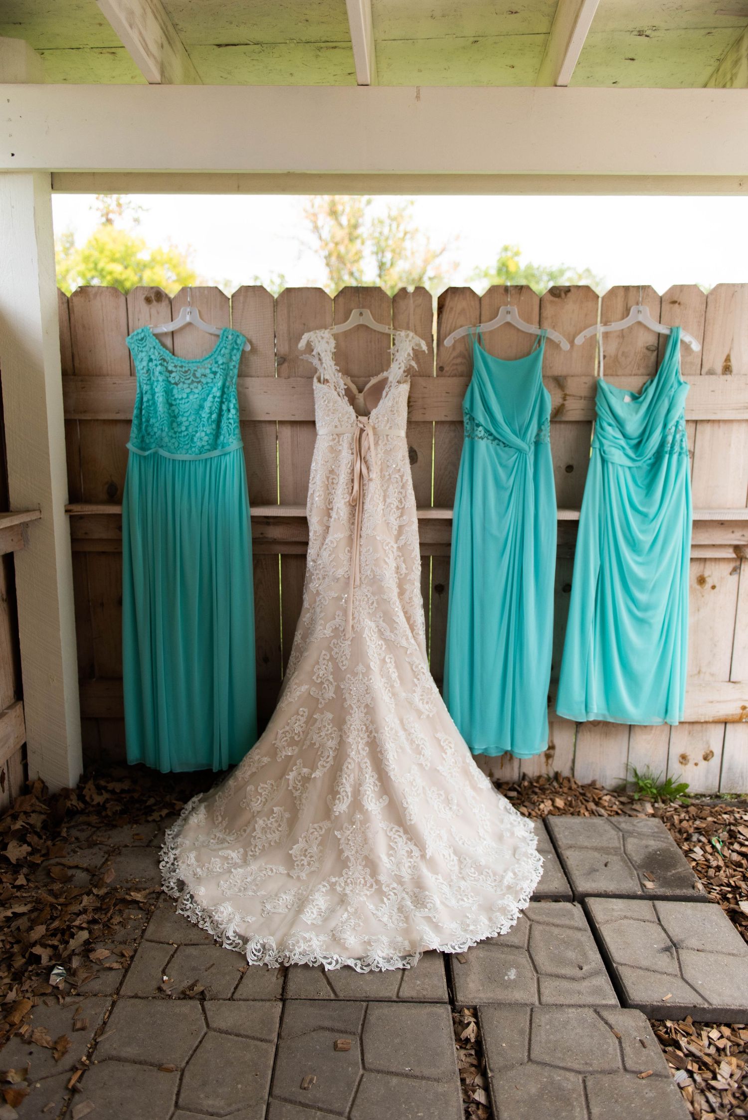 Turquoise bridesmaid dresses hanging beside a white lace wedding gown against a wooden fence.