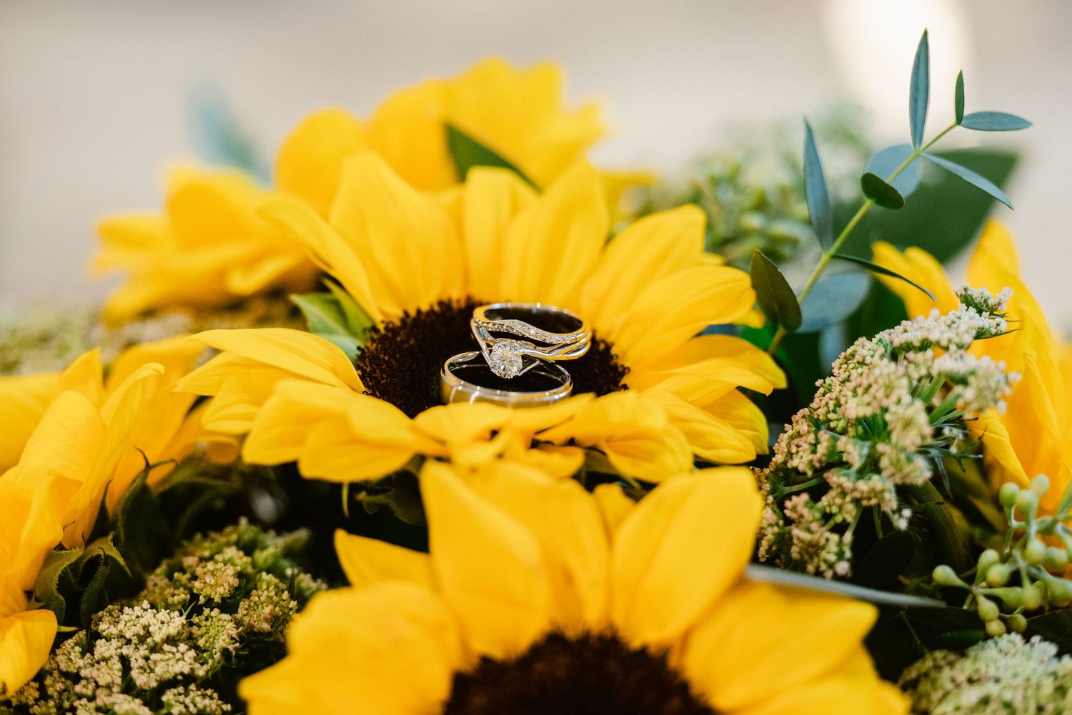 Wedding rings displayed on vibrant yellow sunflower petals with greenery in a wedding bouquet arrangement.