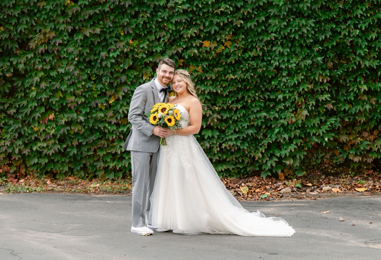 Couple embraces and poses for wedding portrait against lush ivy-covered wall outdoors.