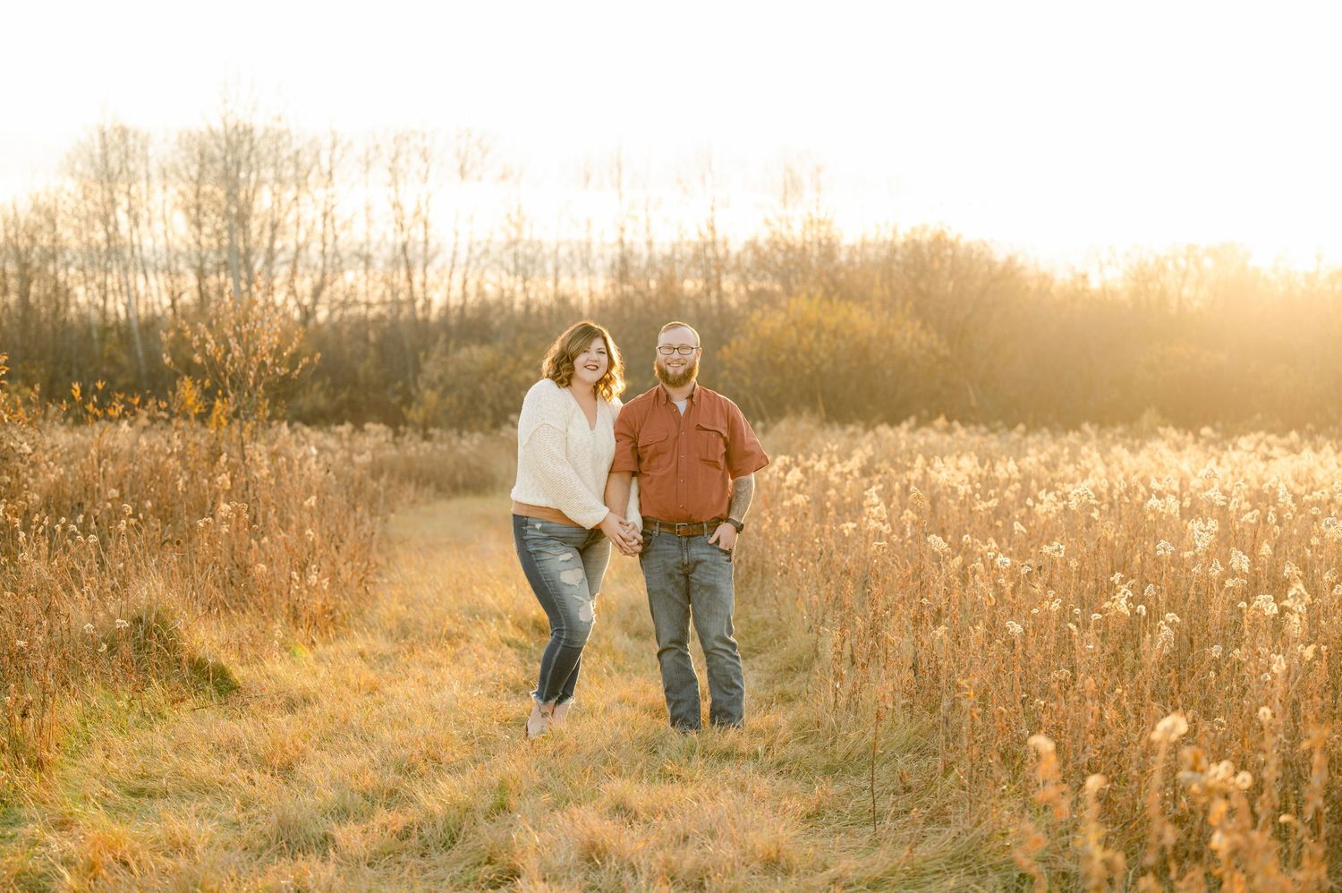 Two people walking together through a golden autumn field during sunset with tall grass and trees in the background. St cloud area engagemen