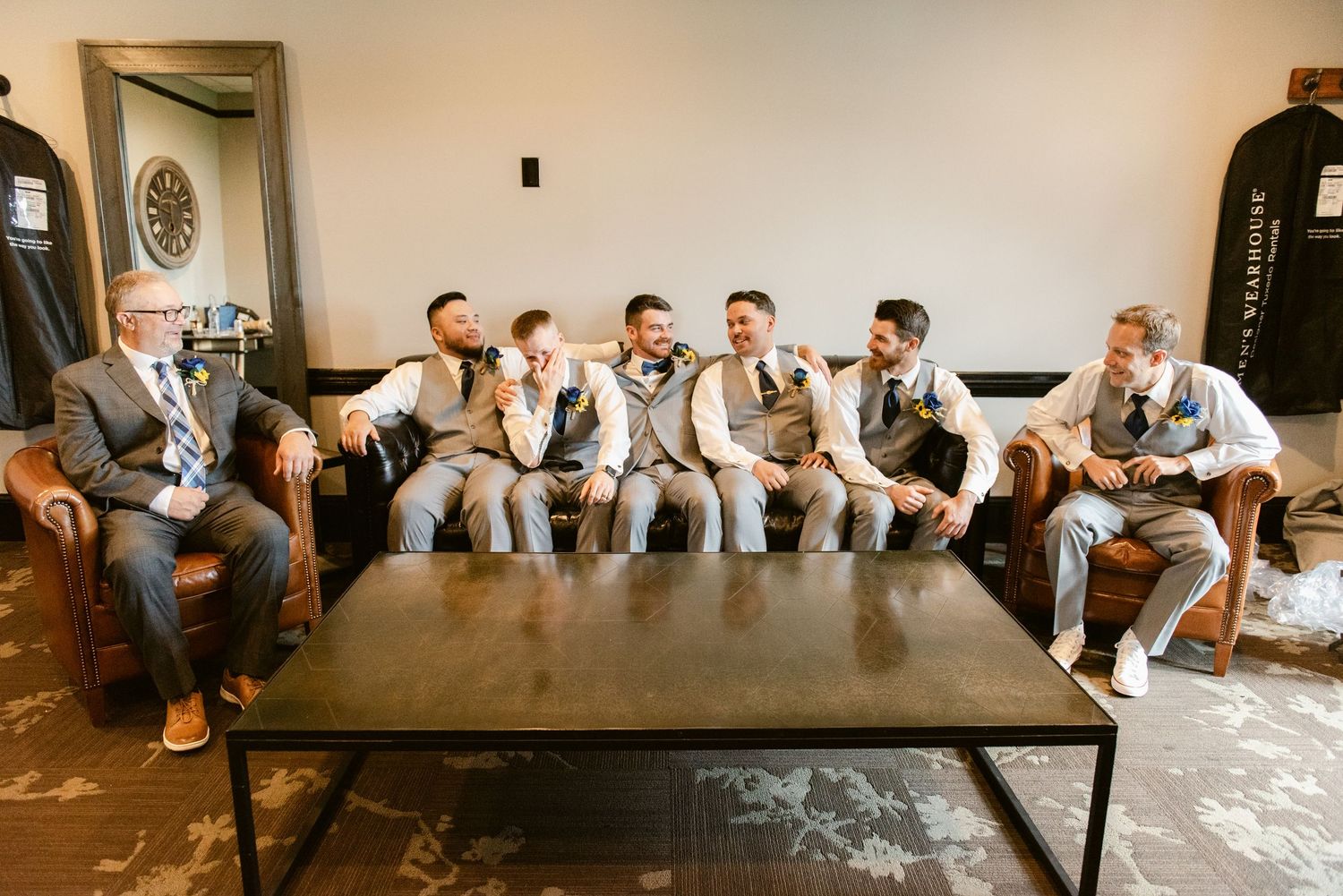 Groomsmen in matching grey vests and pants relax on a leather couch before the wedding.