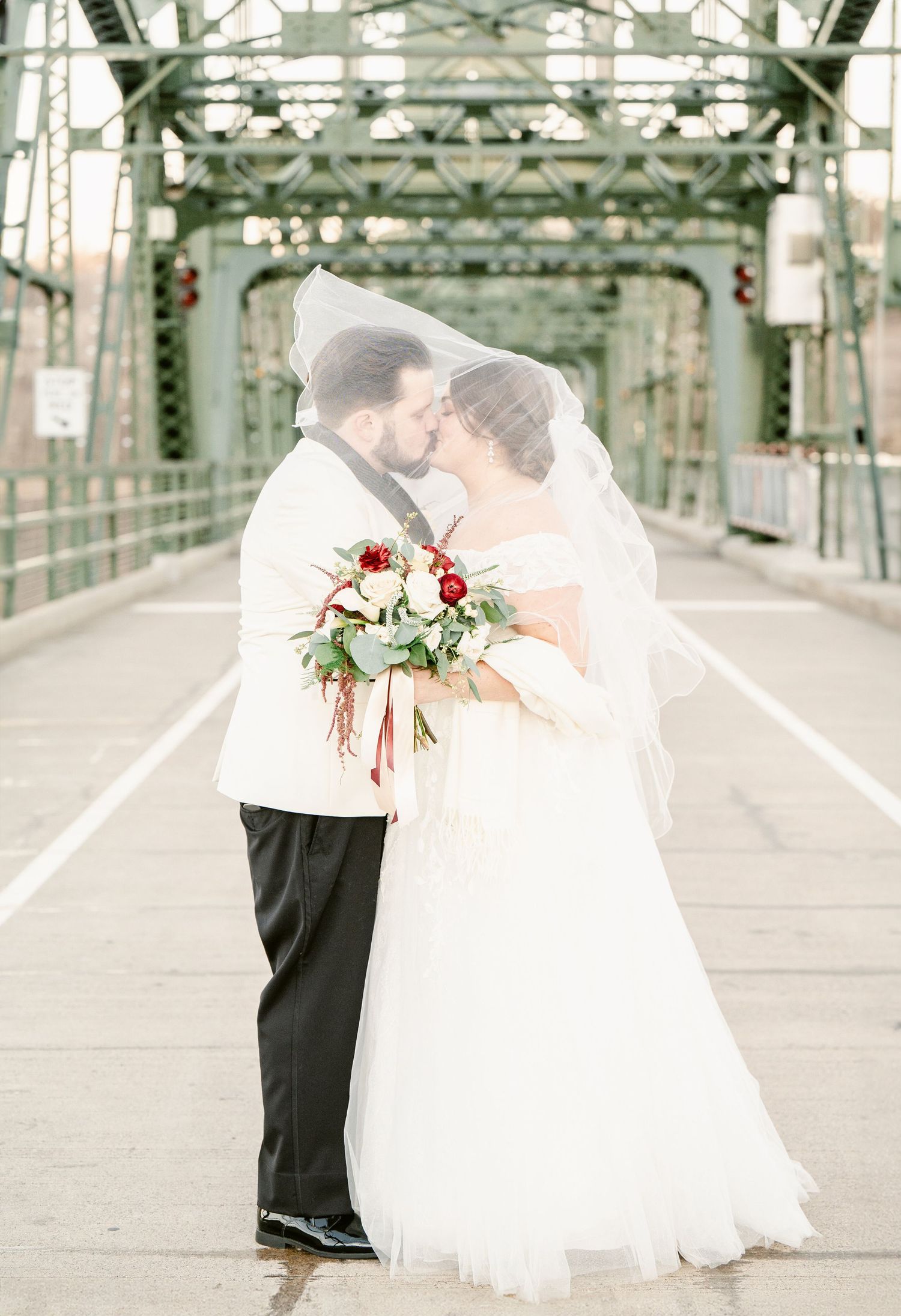 affordable st cloud area. Newlywed couple embraces romantically on an elegant steel bridge while holding a red and white floral bouquet.