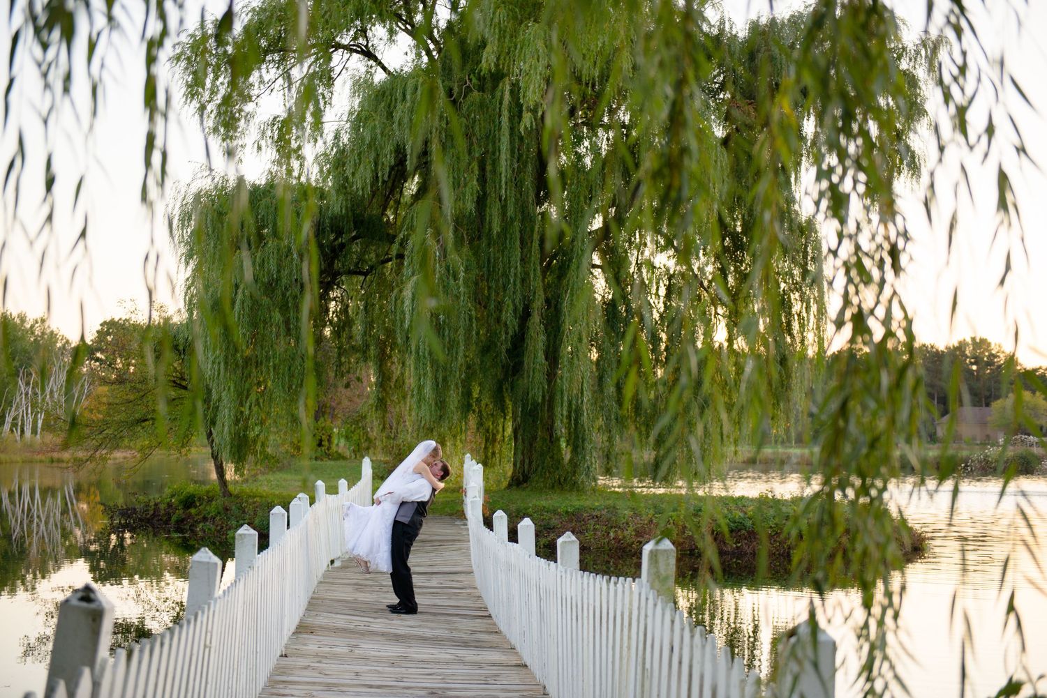 Couple shares romantic moment on white wooden bridge beneath drooping willow branches at sunset. Andover wedding at Magestic Oaks Golf Club