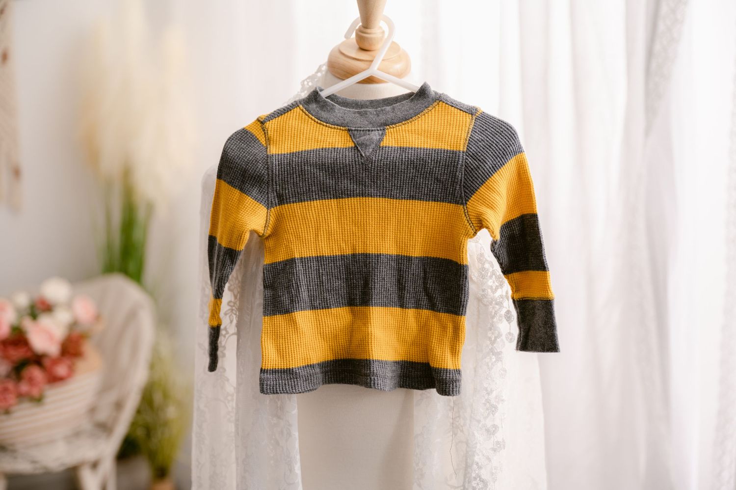 A yellow and gray striped long-sleeve children's shirt hanging on a wooden mannequin bust against a sheer curtain backdrop.