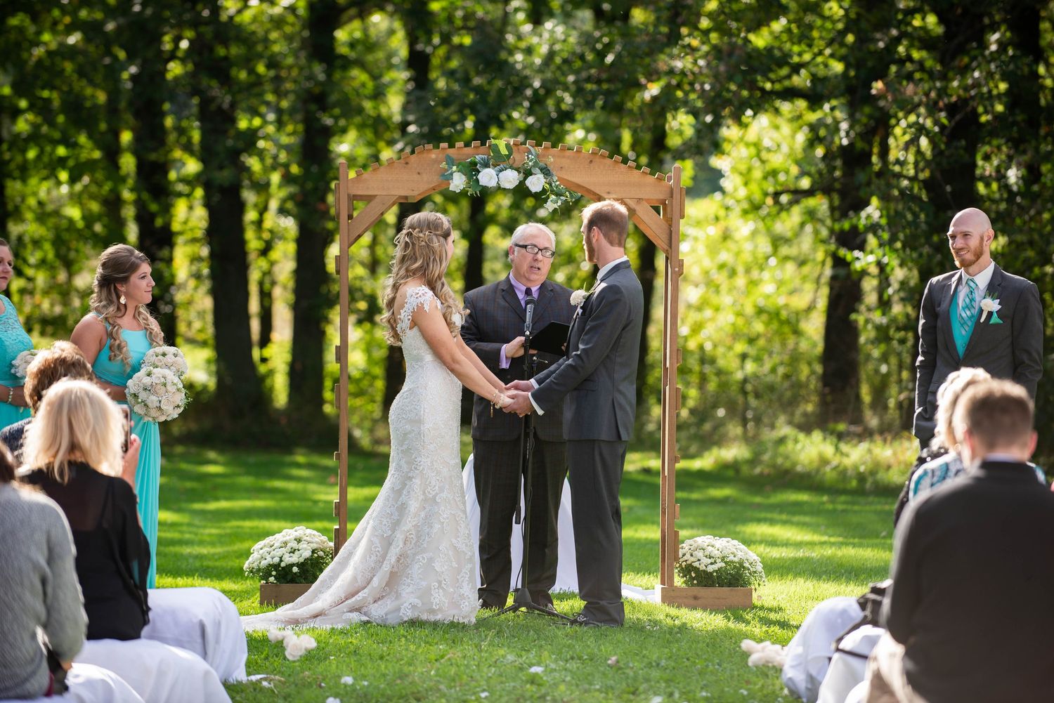 Wedding ceremony takes place in a sunlit wooded park setting with guests seated on white chairs. Rum River Barn Milaca Minnesota Weddings