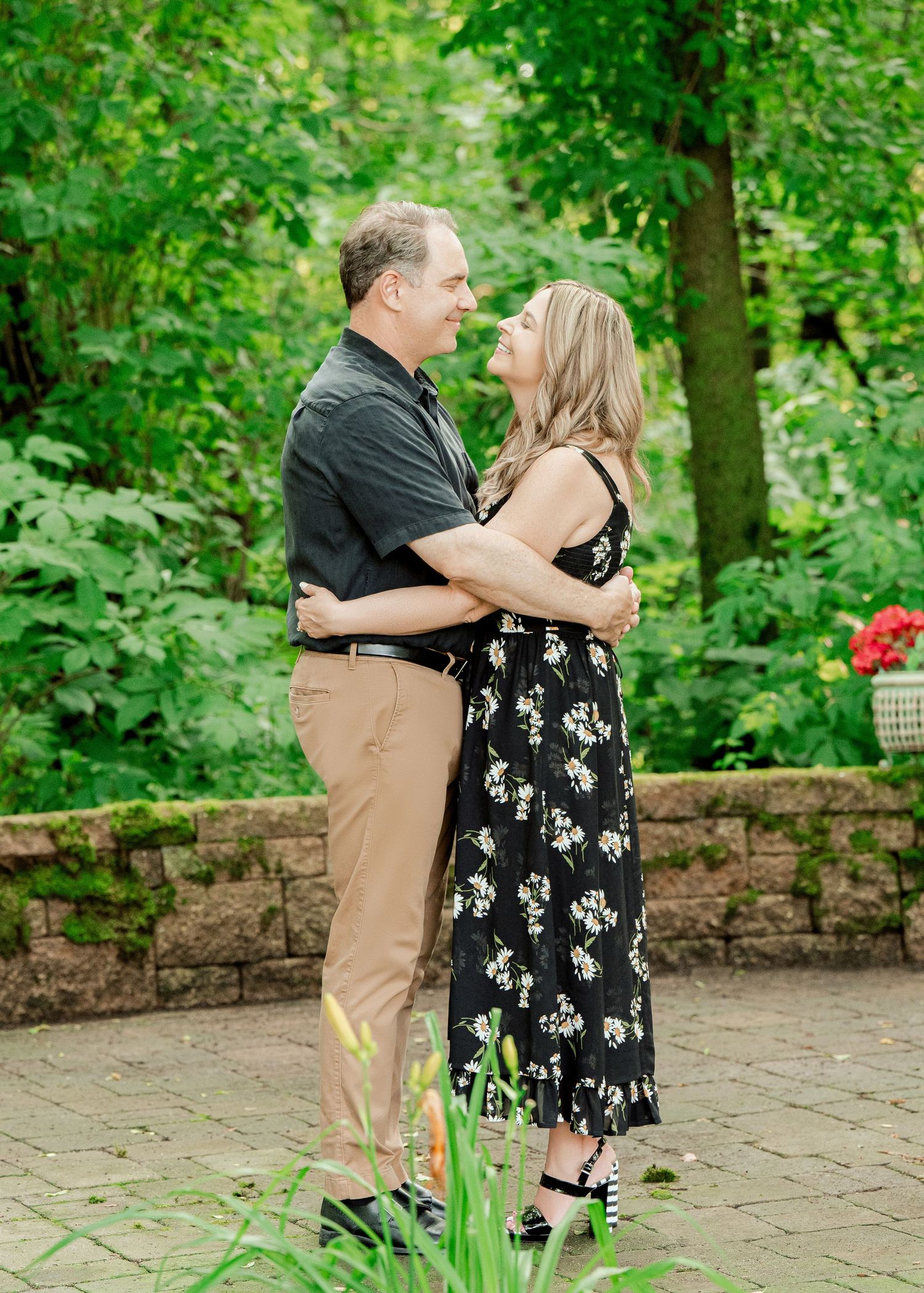 Clear Lake MN Family Photos: A couple embraces on a stone patio surrounded by lush green foliage in a garden setting.