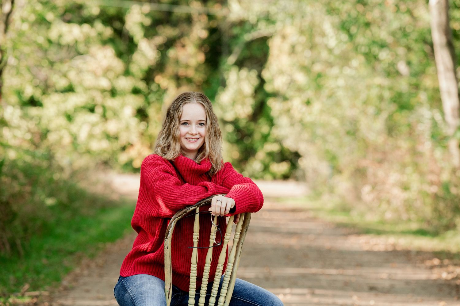 Senior Portrait in Foreston MN in a red sweater and blue jeans sits on a wooden ladder-back chair outdoors among autumn trees.