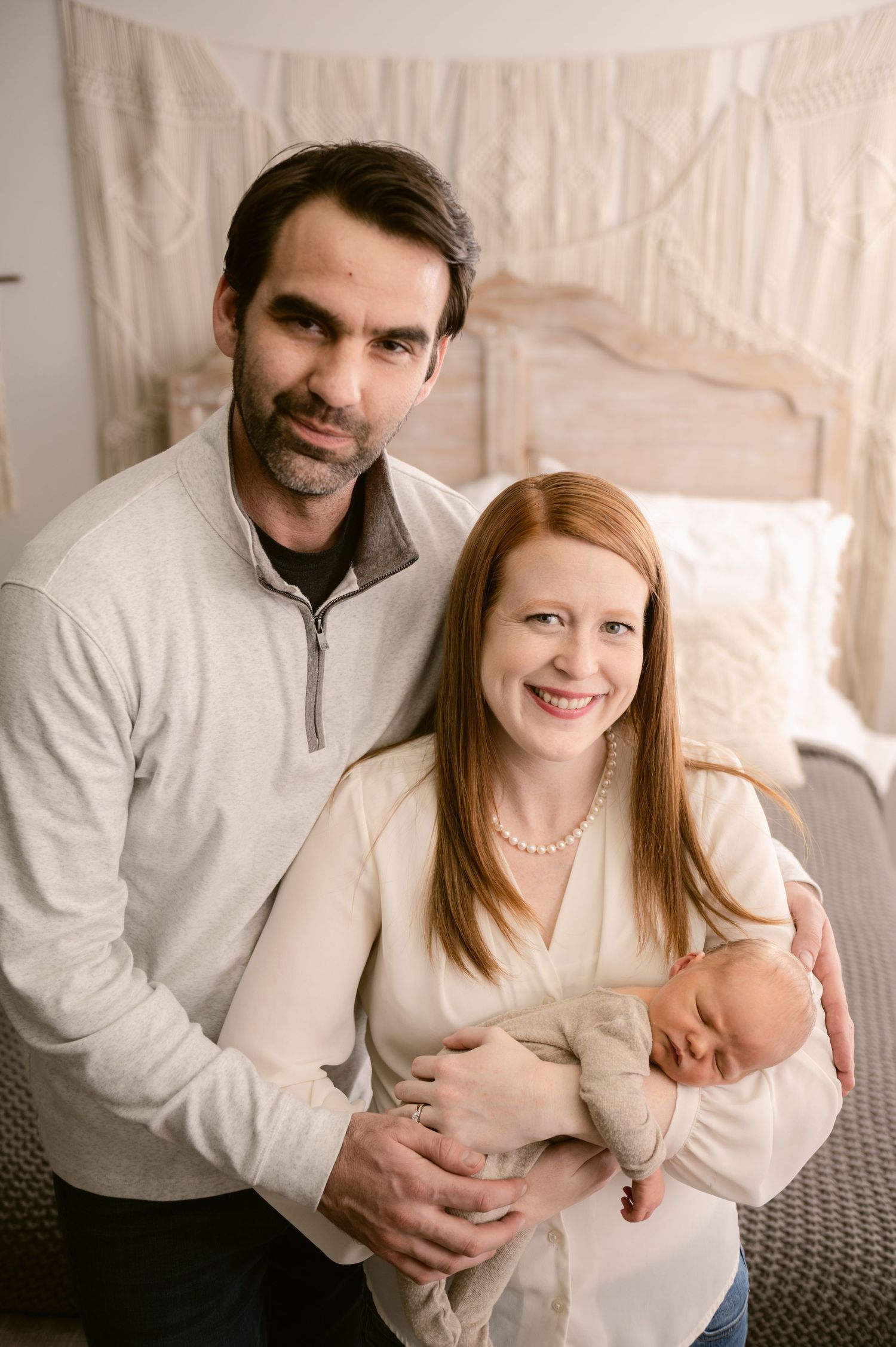 Family of three poses together indoors wearing cream colored outfits with newborn baby.