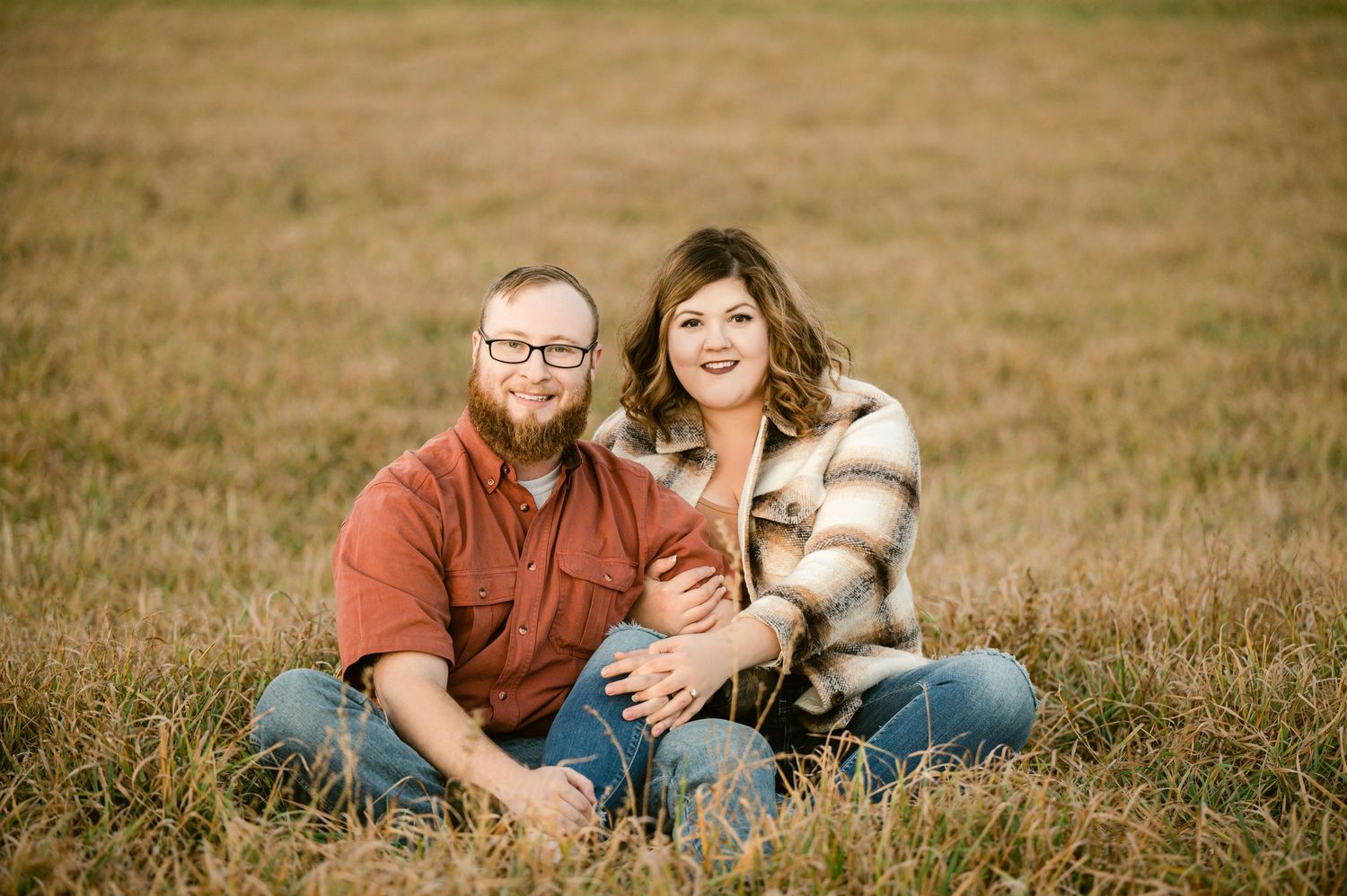 A couple sits together in a grassy field on an autumn day wearing casual attire and smiling at the camera. Princeton MN Milestone, Maternity