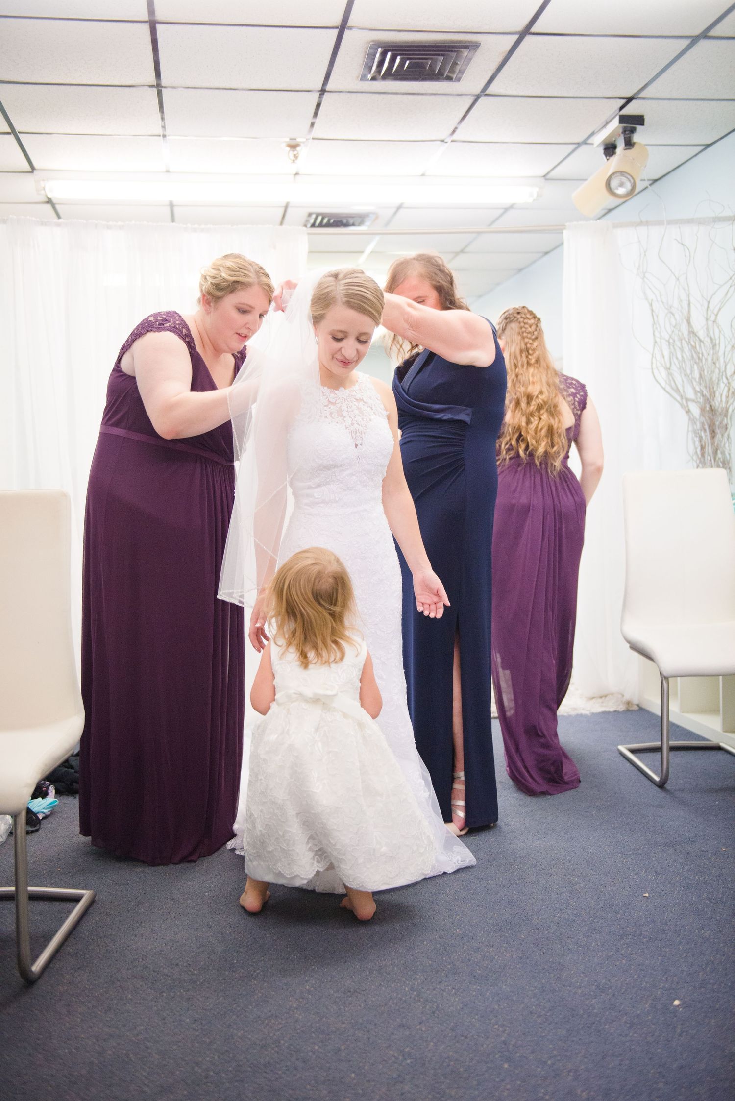 Wedding party helps adjust a white lace wedding gown while a small child in white looks on in a dressing room. Andover wedding at Magestic O