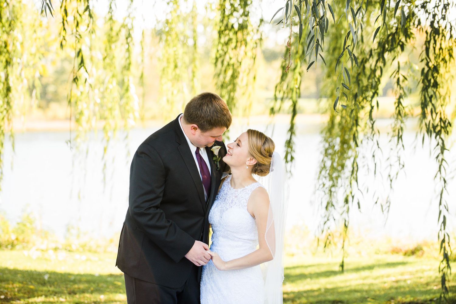 Elegant wedding couple shares a romantic moment under a weeping willow tree by a sunlit lake. Andover wedding at Magestic Oaks Golf Club