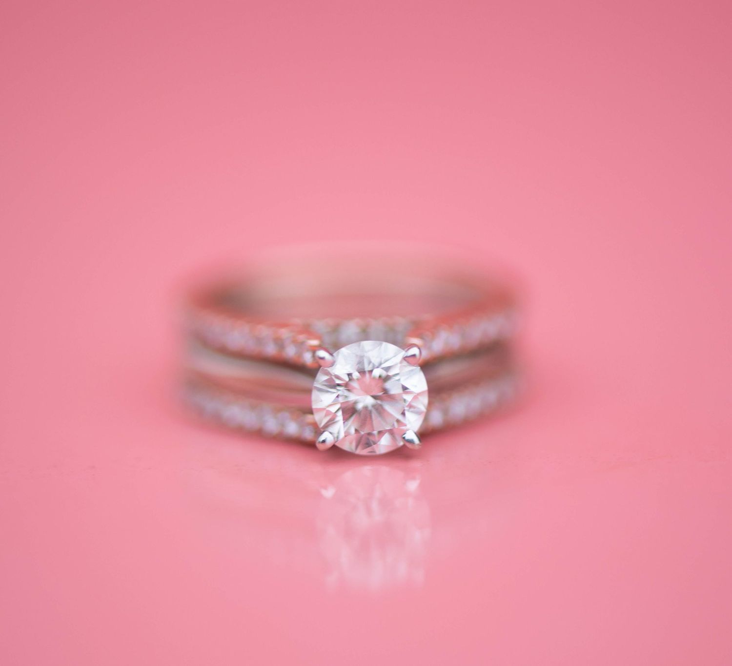 A delicate solitaire engagement ring with a round diamond sparkles against a soft pink background.