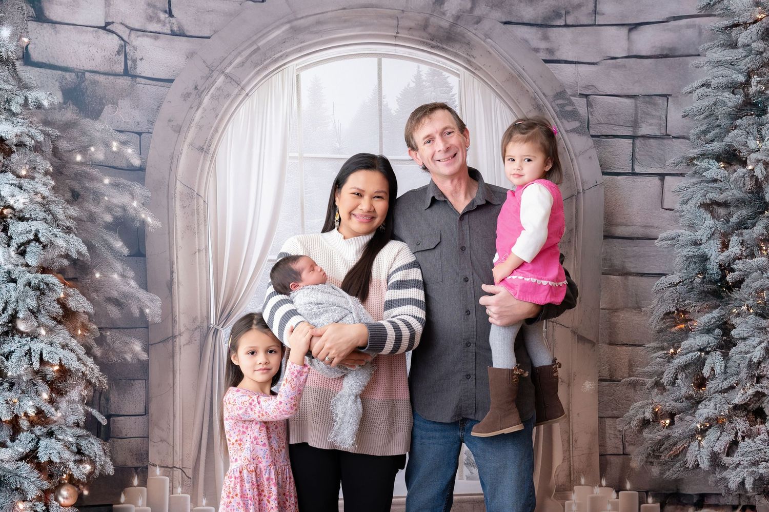 Family poses under stone arch with winter decorations and snow-covered trees in professional studio portrait setting.