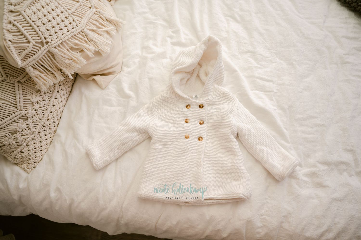 A cozy white knit baby sweater with gold buttons lies on a cream colored textured bedspread.
