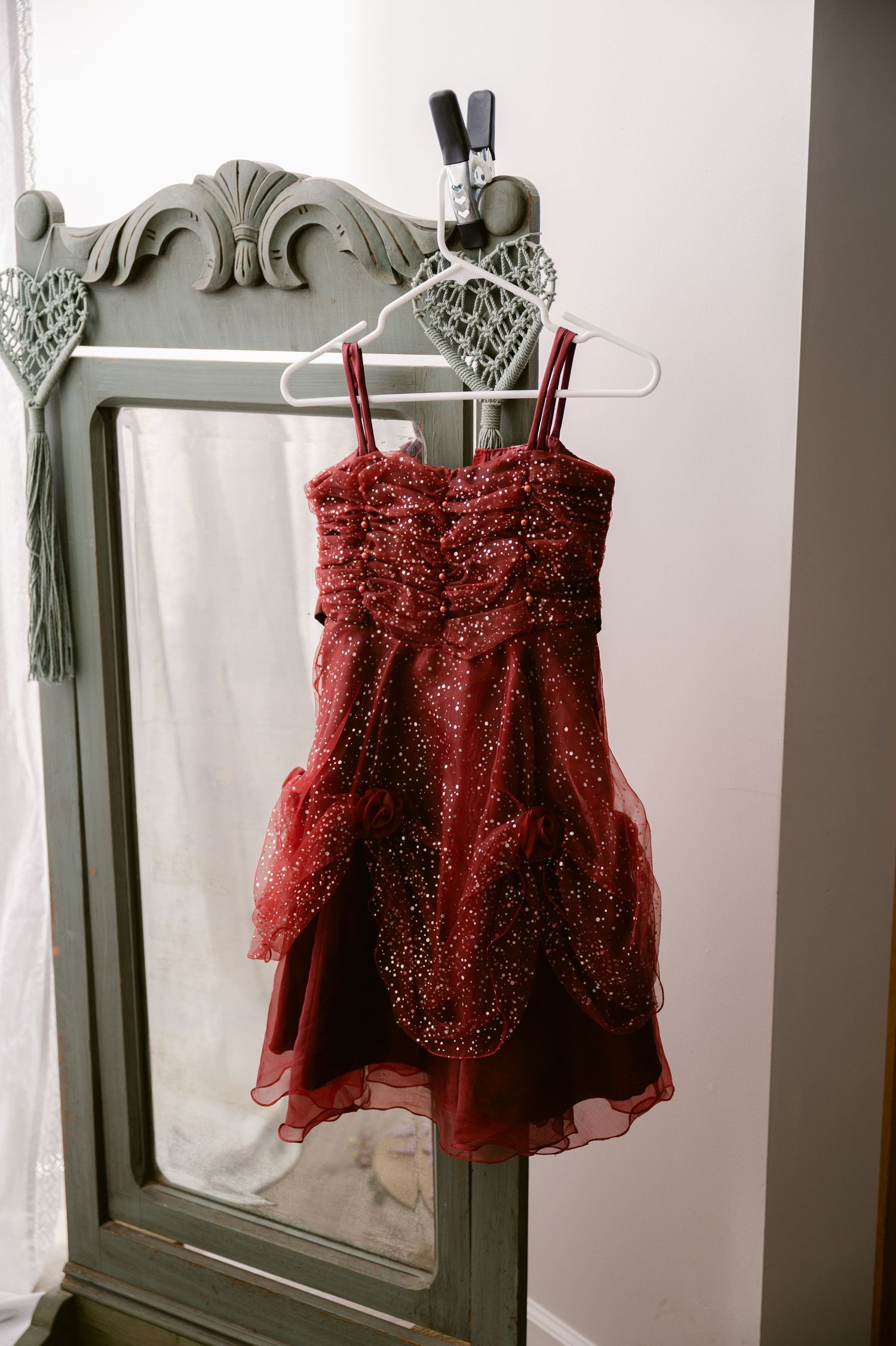 A sparkly red formal dress with ruched bodice hangs on a white hanger against a vintage gray framed mirror.