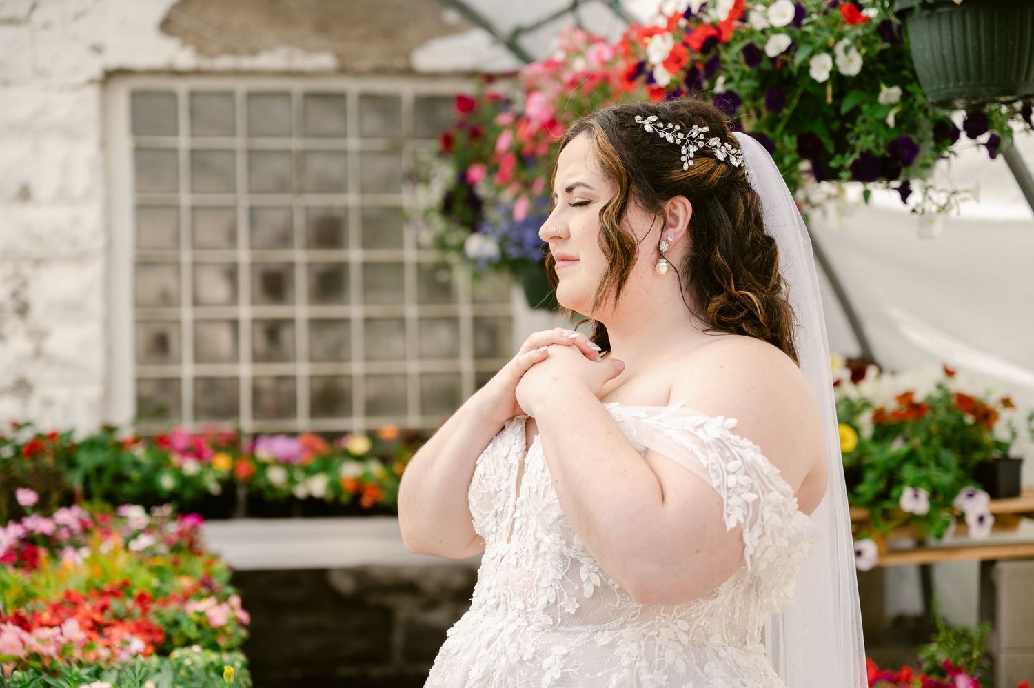 Bride in lace gown poses by colorful flower garden on wedding day.