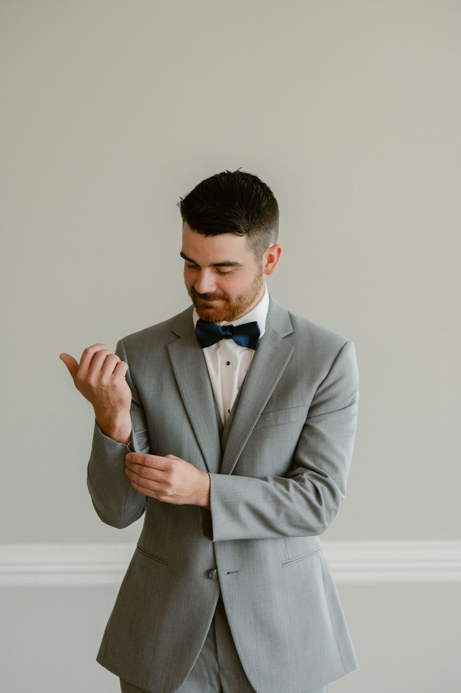 A person in a grey suit and black bow tie adjusts their sleeve cuff against a neutral background.