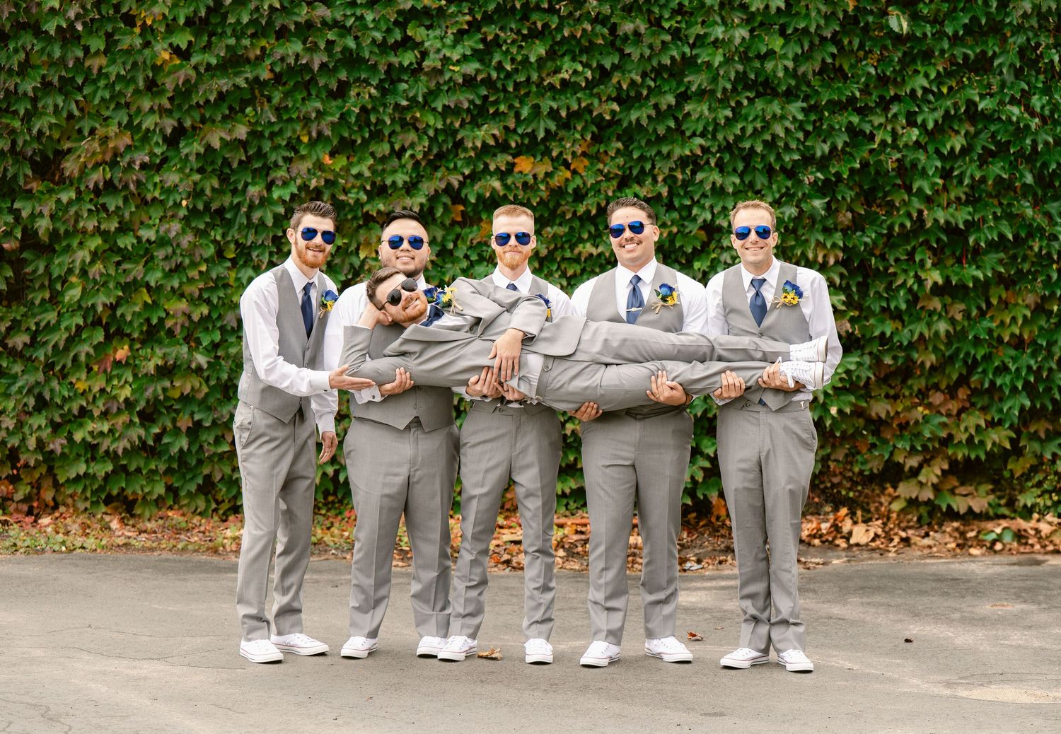 Groomsmen in matching grey suits and sneakers pose against an ivy-covered wall.